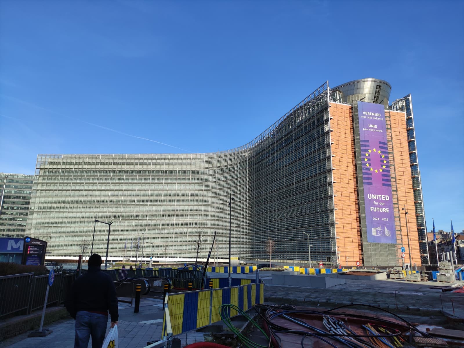 Ein großes modernes Gebäude mit einer geschwungenen Glasfassade, auf der ein großes lila Banner mit der EU-Flagge und der Aufschrift 'United for the Future' zu sehen ist. Im Vordergrund sind Baumaterialien und eine Person im Gehen zu sehen.