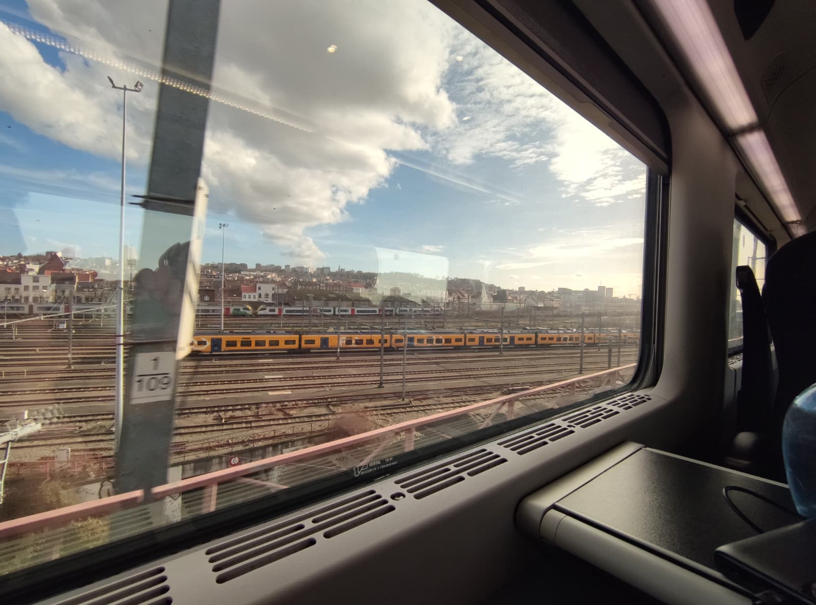 Blick aus dem Inneren eines Zuges durch ein großes Fenster auf Eisenbahnschienen, einen vorbeifahrenden gelben Zug und eine Stadtlandschaft unter einem teilweise bewölkten Himmel. Im Vordergrund sind Zugsitze und ein Tisch zu sehen.