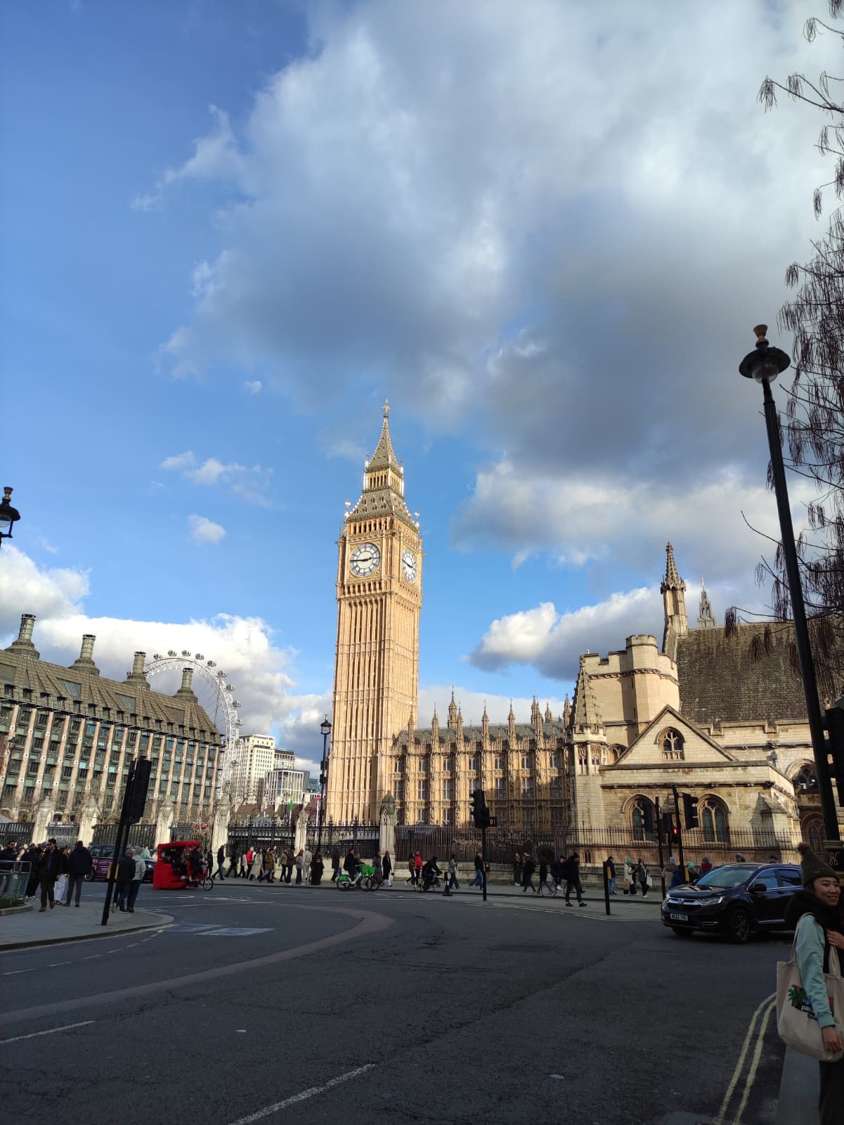Das Foto zeigt den Londoner Uhrenturm Big Ben unter einem blauen Himmel mit weißen Wolken. Im Vordergrund sind Menschen und Autos auf der Straße zu sehen, im Hintergrund historische Gebäude und das London Eye.