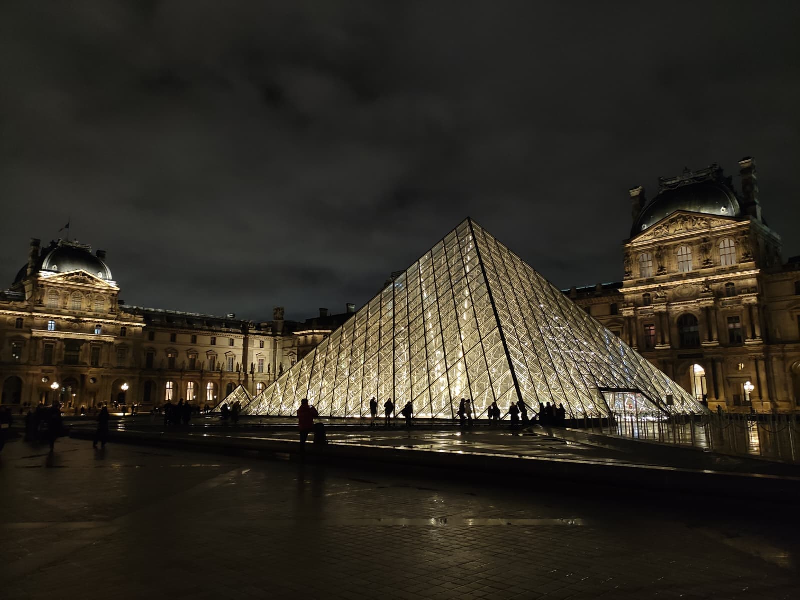 Die beleuchtete Glaspyramide des Louvre-Museums in Paris bei Nacht, umgeben von historischen Gebäuden unter einem wolkenverhangenen Himmel, mit Silhouetten von Menschen rund um das Gebäude.