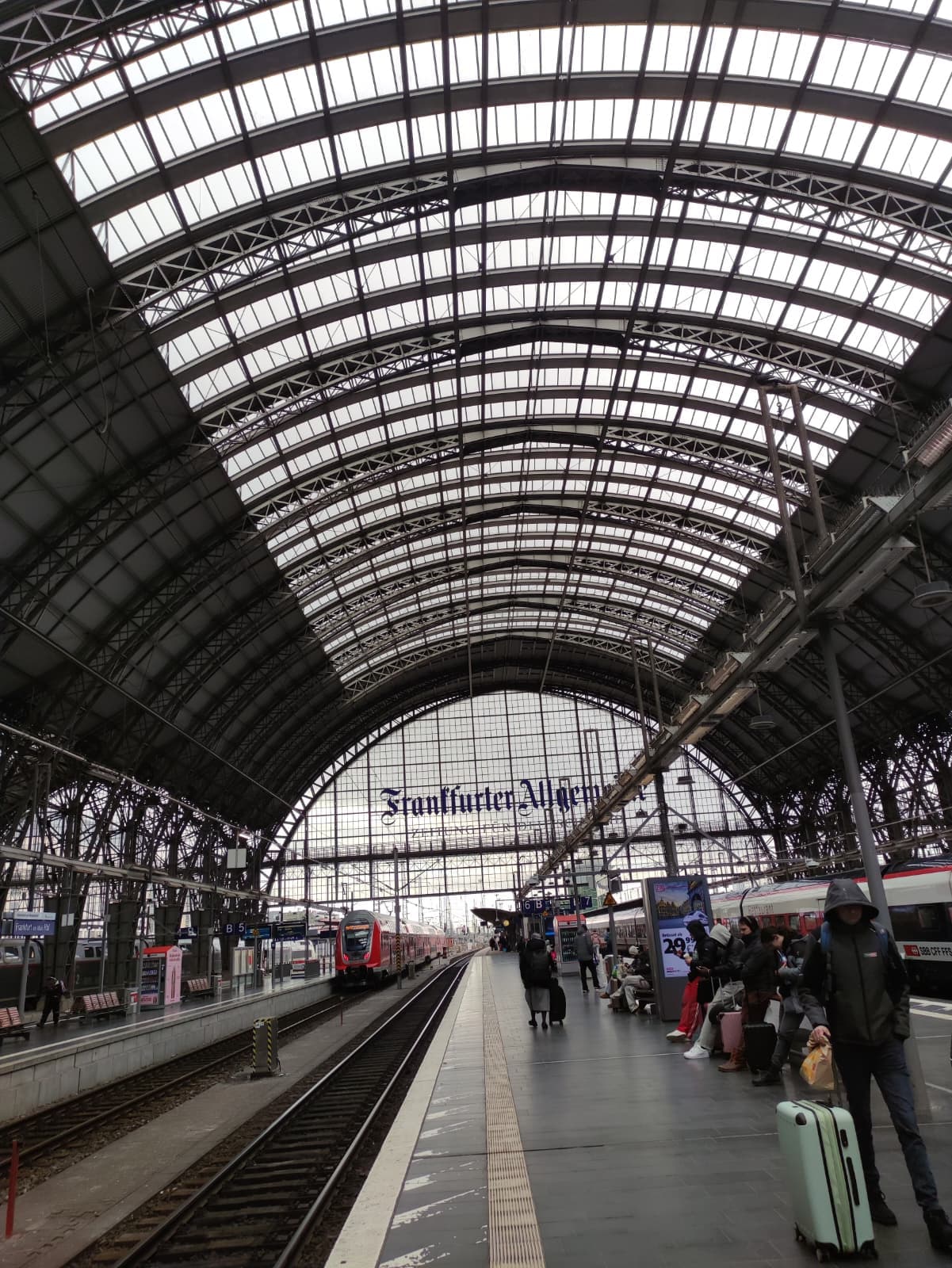 Menschen warten mit Gepäck in einem belebten Bahnhof unter einem großen gewölbten Dach aus Glas und Stahl. Auf den Gleisen fahren Züge, und im Hintergrund steht ein Schild mit der Aufschrift 'Frankfurter'. Die Atmosphäre ist hektisch und bewölkt.