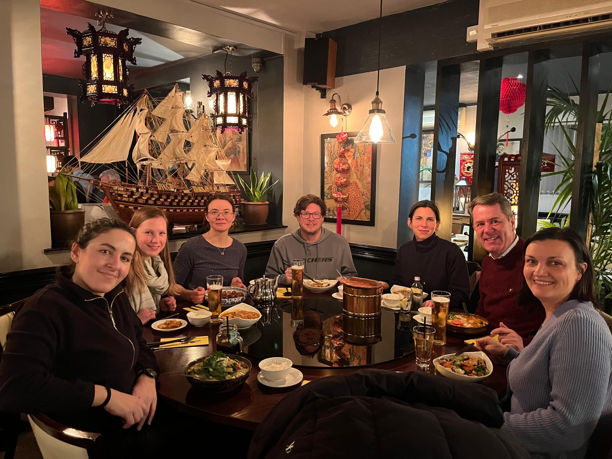 Sieben Personen sitzen um einen runden Tisch in einem warm beleuchteten Restaurant und teilen sich eine Mahlzeit mit verschiedenen Speisen und Getränken. Im Hintergrund sind dekorative Laternen, Pflanzen und ein großes Modellschiff zu sehen.