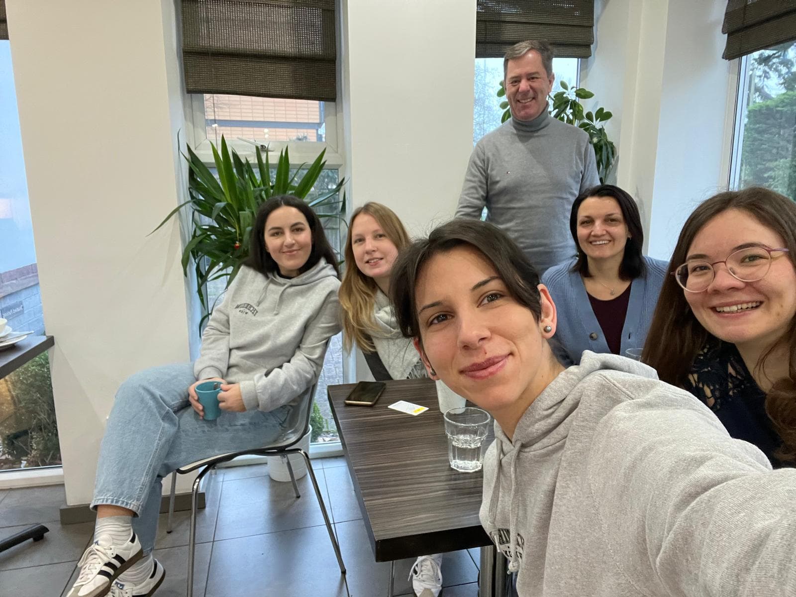 Fünf Personen, vier Frauen und ein Mann, lächeln und posieren für ein Selfie in einem hellen Innenraum mit großen Fenstern, Grünpflanzen und einem Tisch mit einem Glas Wasser und einem Telefon.