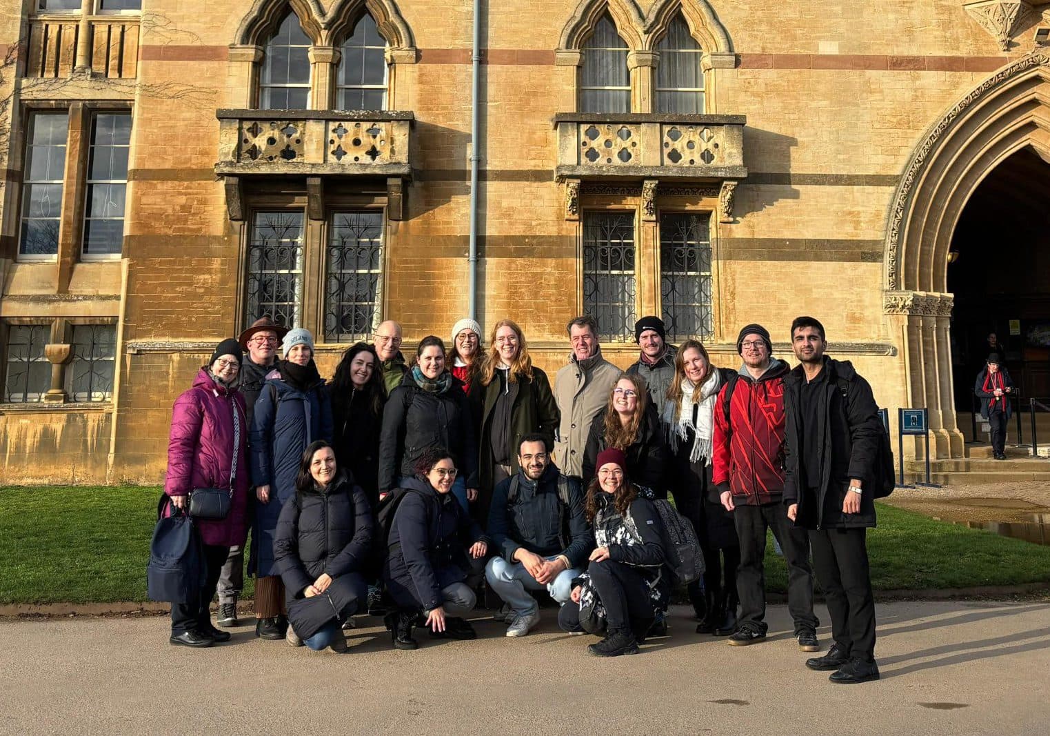 Eine Gruppe von Menschen posiert an einem sonnigen Tag für ein Foto vor einem großen historischen Steingebäude mit Rundbogenfenstern und einem bogenförmigen Eingang.
