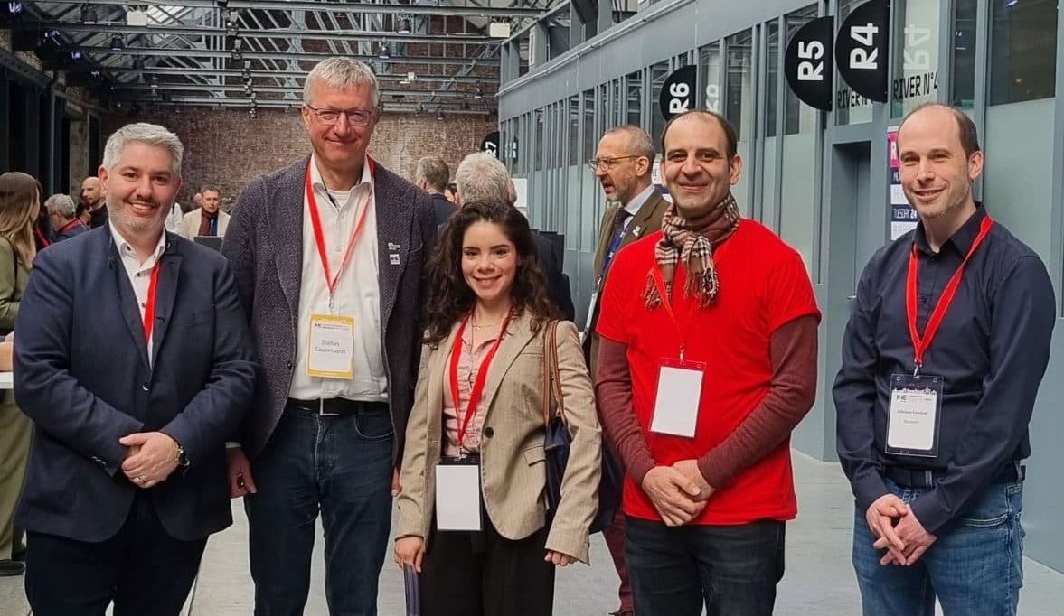 Fünf Personen, die Konferenzausweise und rote Schlüsselbänder tragen, stehen für ein Gruppenfoto in einer Halle im industriellen Stil mit Glasdecken und sichtbaren Balken. Andere Teilnehmer sind im Hintergrund zu sehen.
