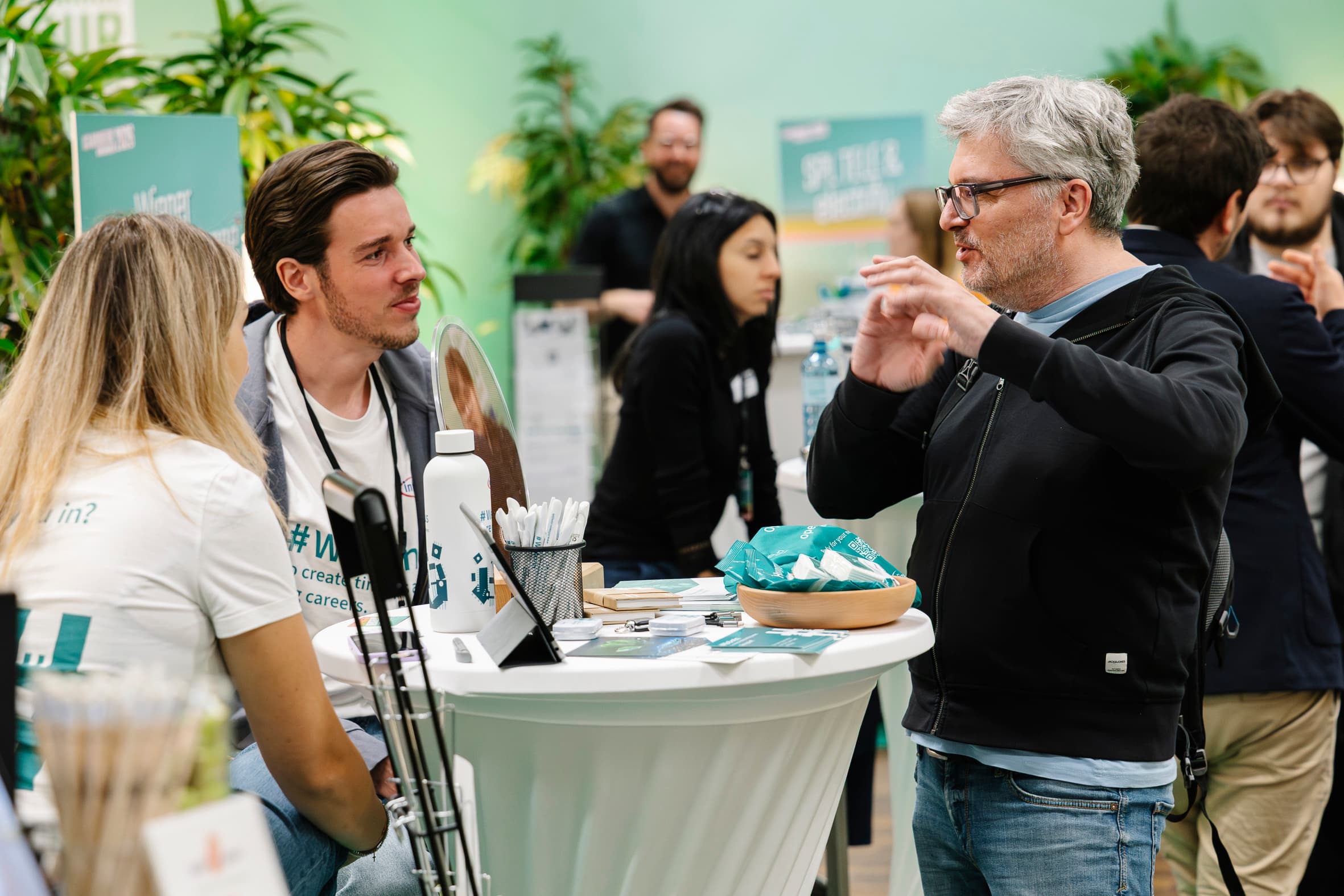 Bei einer Indoor-Veranstaltung stehen und sitzen mehrere Personen um einen hohen runden Tisch herum und unterhalten sich. Auf dem Tisch sind Werbeartikel und Informationsmaterial zu sehen. Der Hintergrund besteht aus Topfpflanzen und Veranstaltungsschildern.