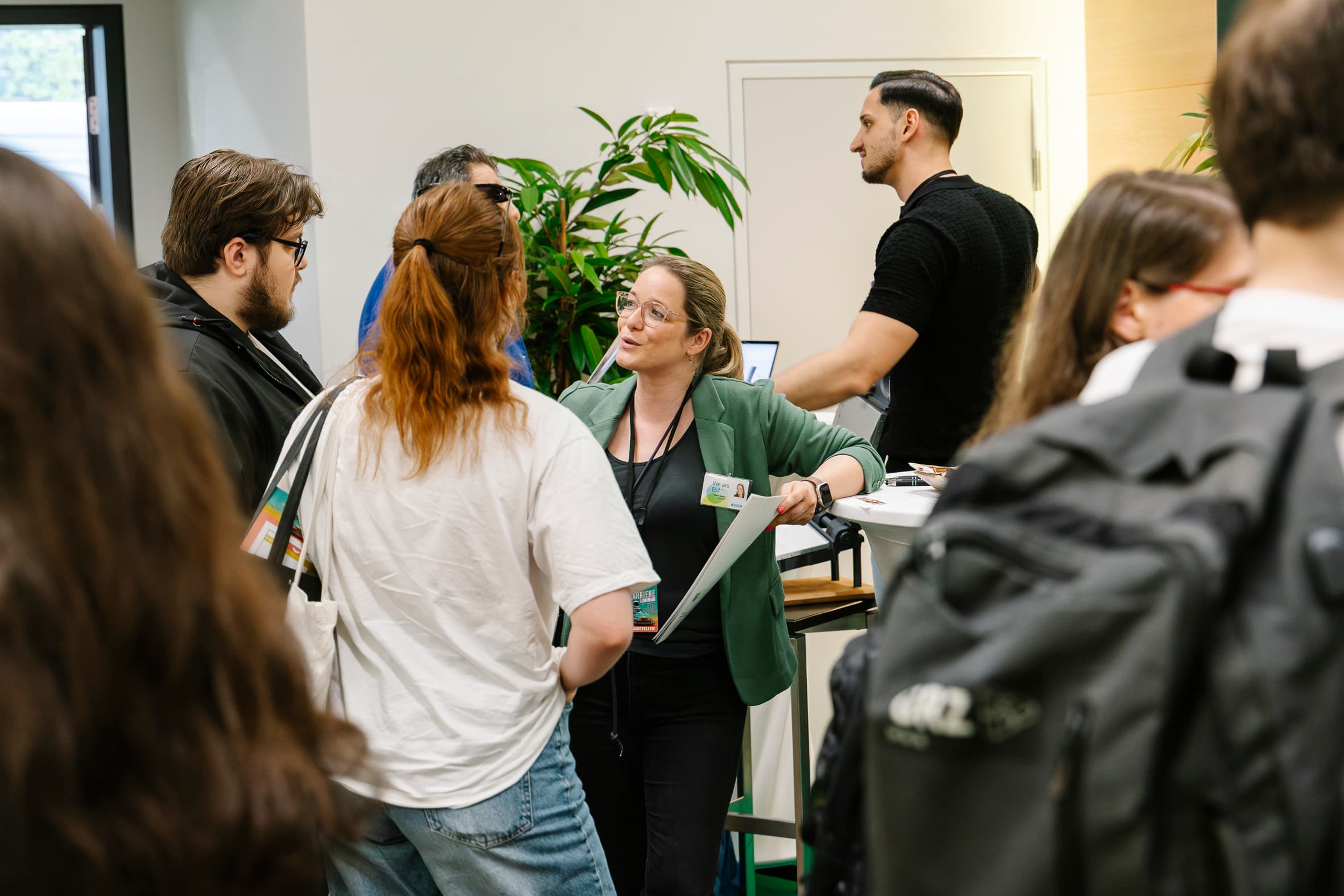 Eine Gruppe von Menschen steht und unterhält sich bei einer Indoor-Veranstaltung. Eine Frau in einem grünen Blazer spricht zu anderen in der Mitte. Einige Teilnehmer tragen Abzeichen und Rucksäcke, und der Hintergrund ist mit Grünpflanzen geschmückt.