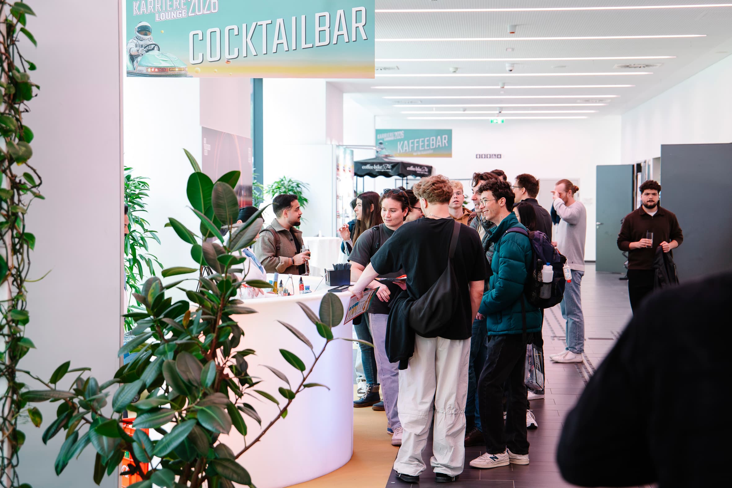 Die Menschen sind an einer weißen Cocktailbar in einem modernen Innenraum versammelt, mit Pflanzen in der Nähe und einem 'COCKTAILBAR'-Schild darüber. Einige Leute bestellen Getränke, während andere sich unterhalten. Die Atmosphäre wirkt ungezwungen und lebendig.