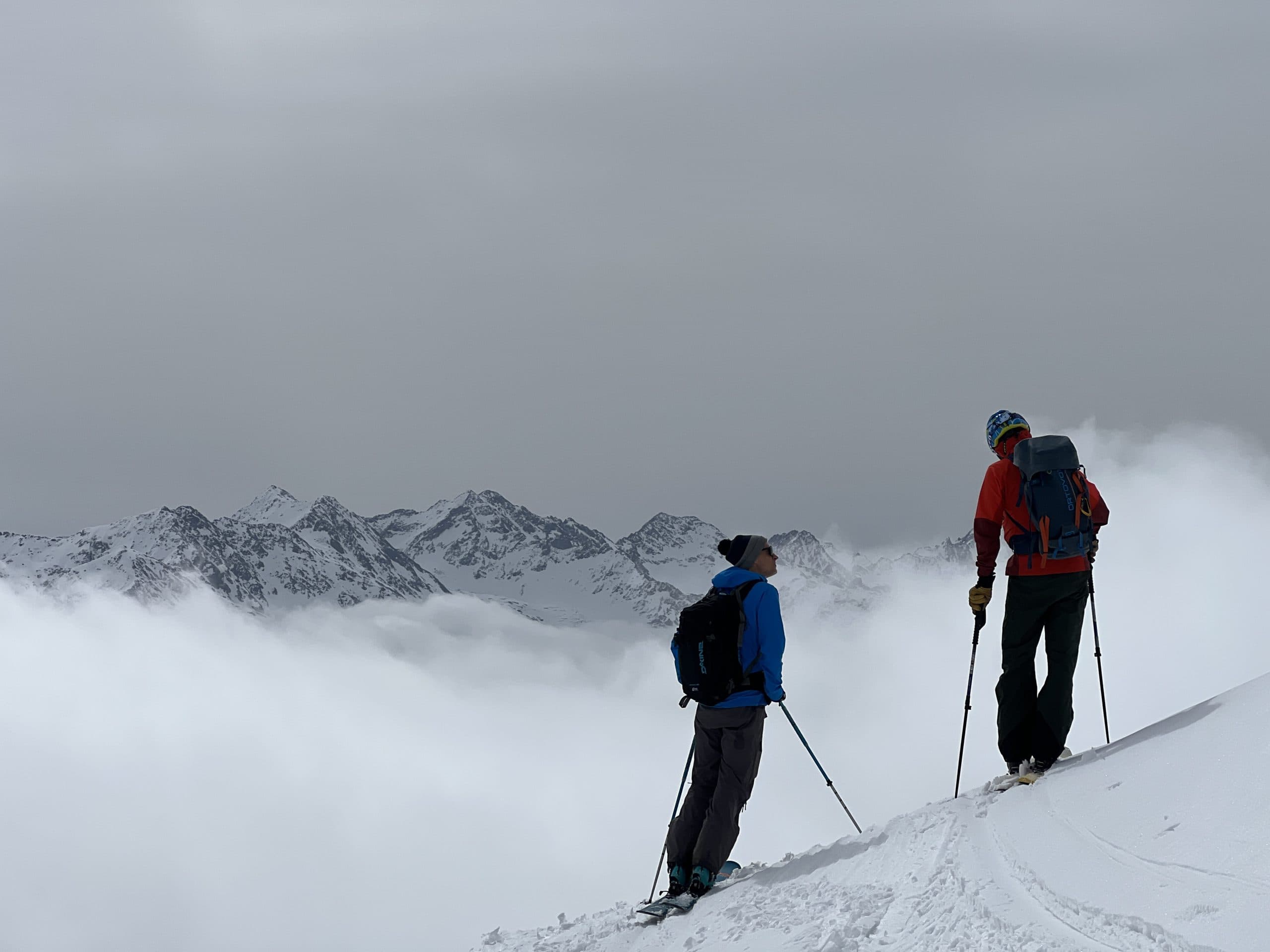 Zwei Skifahrer stehen an einem verschneiten Berghang und blicken auf ferne schneebedeckte Gipfel unter einem bewölkten Himmel. Dicke Wolken füllen das Tal unter ihnen und verdecken teilweise die niedrigeren Berge.