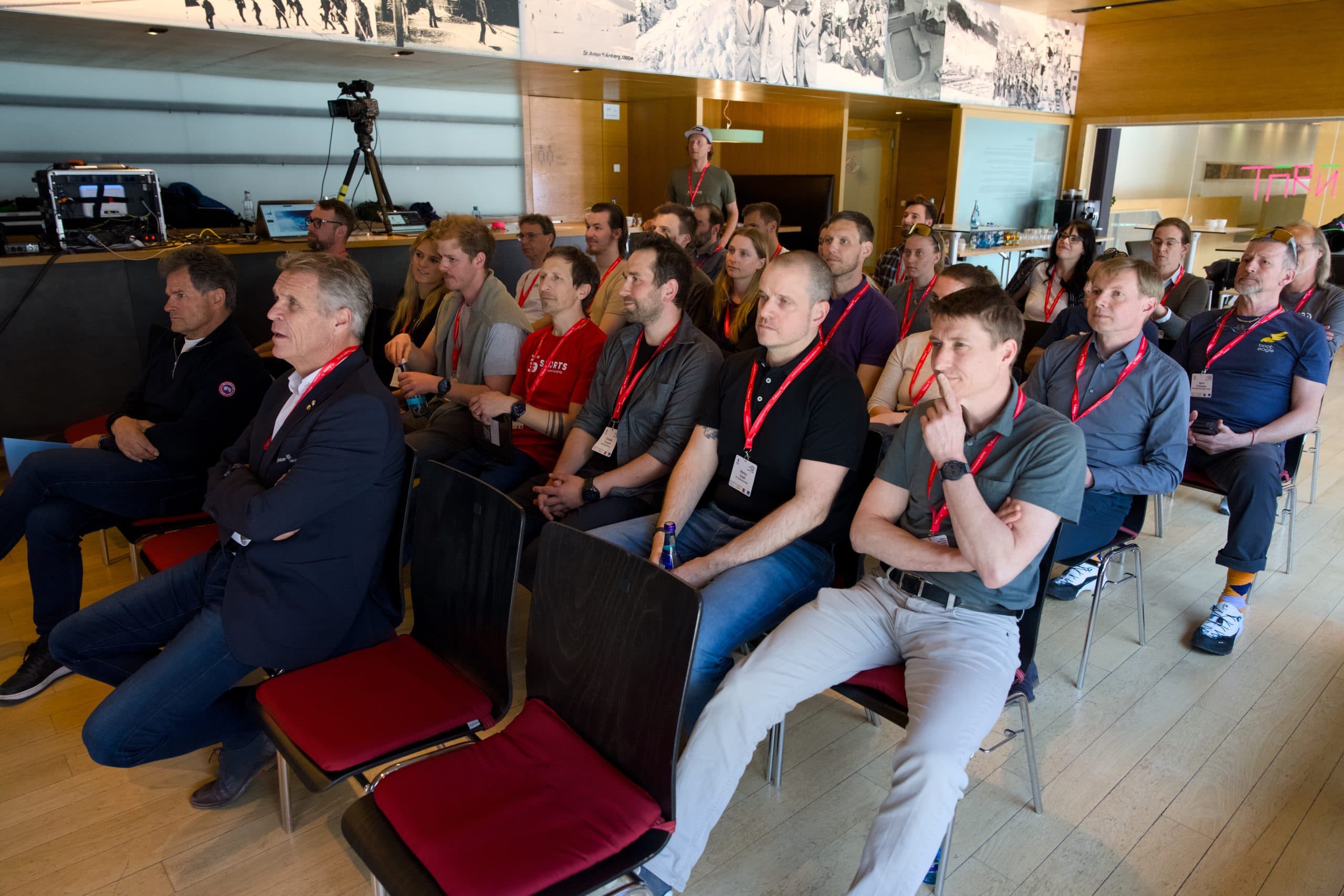 Eine Gruppe von Personen mit roten Schlüsselbändern sitzt aufmerksam in Stuhlreihen bei einer Veranstaltung oder einem Seminar in einer Halle. Einige haben die Arme verschränkt, und einige lehnen sich nach vorne und konzentrieren sich auf einen Redner außerhalb des Bildes.