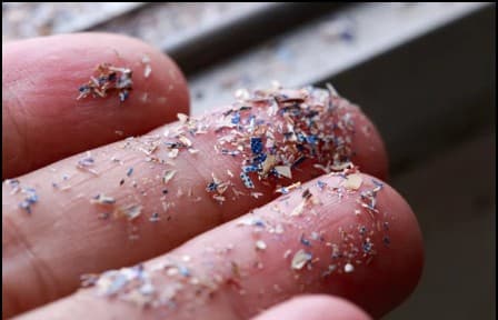 Die Finger sind mit winzigen, bunten Mikroplastikpartikeln und -fragmenten bedeckt, die auf Umweltverschmutzung und Umweltprobleme hinweisen. Der Hintergrund ist unscharf und lenkt die Aufmerksamkeit auf die Partikel auf der Haut.
