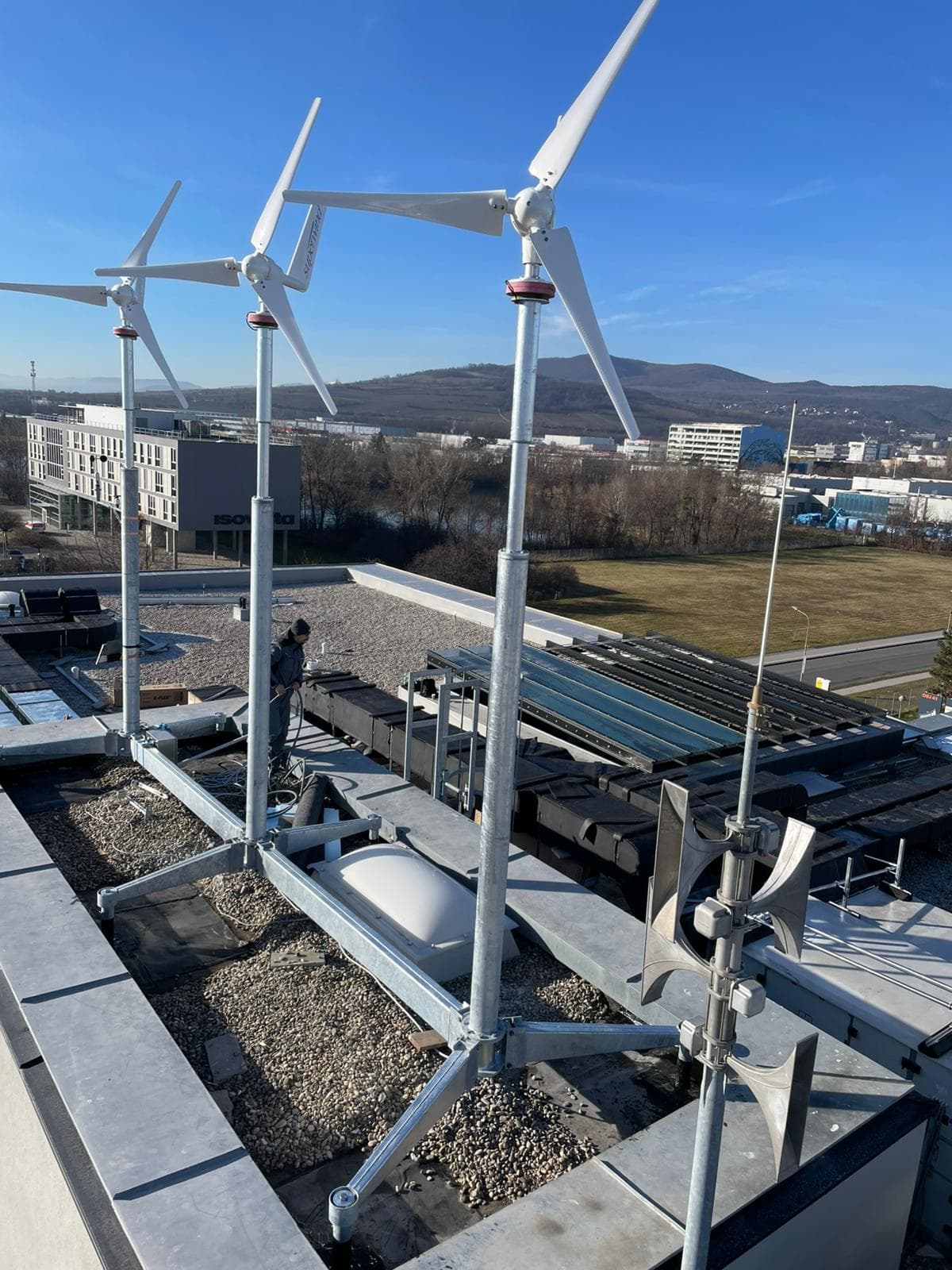 Auf einem flachen Dach installierte Windturbinen mit Sonnenkollektoren in der Nähe; eine Person steht neben den Anlagen. Im Hintergrund sind städtische Gebäude und Hügel unter einem klaren blauen Himmel zu sehen.