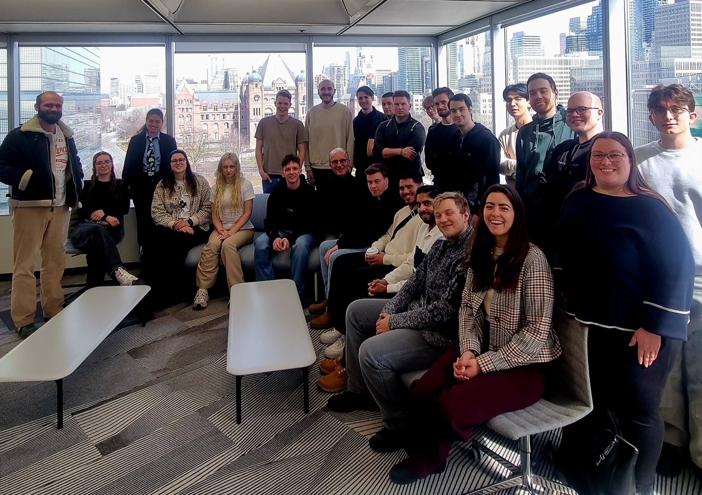 Eine Gruppe von etwa 25 Personen posiert und lächelt für ein Foto in einem modernen, sonnendurchfluteten Büro mit großen Fenstern und einer Stadtlandschaft im Hintergrund. Einige sitzen, während andere hinter ihnen stehen.