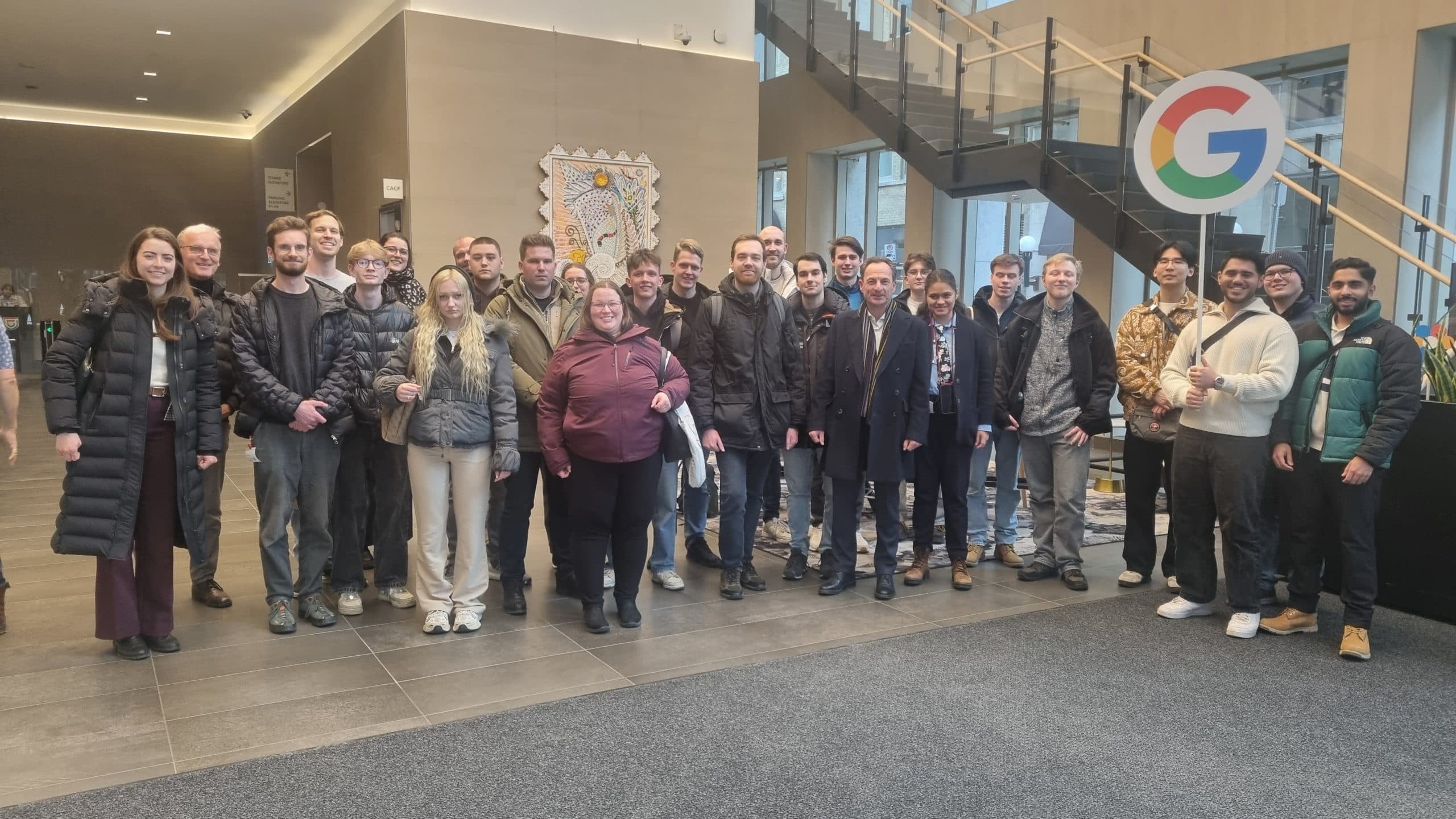 Eine Gruppe von etwa 25 Personen steht und lächelt für ein Foto in einem Bürogebäude neben einem großen Google-Logo und einer Treppe. Die meisten tragen Winterkleidung und stehen auf einem grau gekachelten Boden.