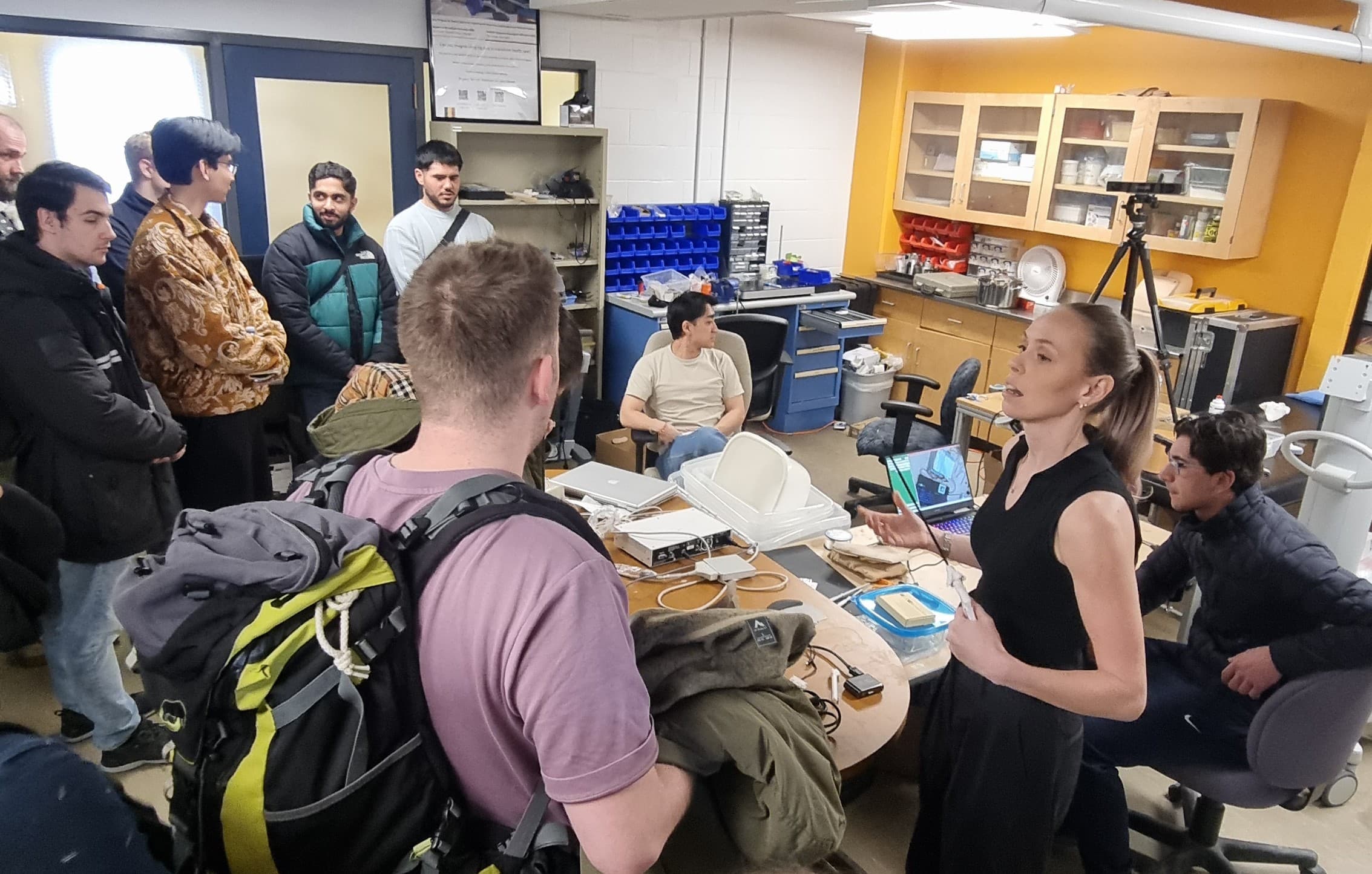 Eine Frau spricht zu einer Gruppe von Menschen in einem geschäftigen Labor voller Geräte, Werkzeuge und Elektronik. Einige Leute sitzen, während andere, die Rucksäcke und Jacken tragen, stehen und aufmerksam zuhören.