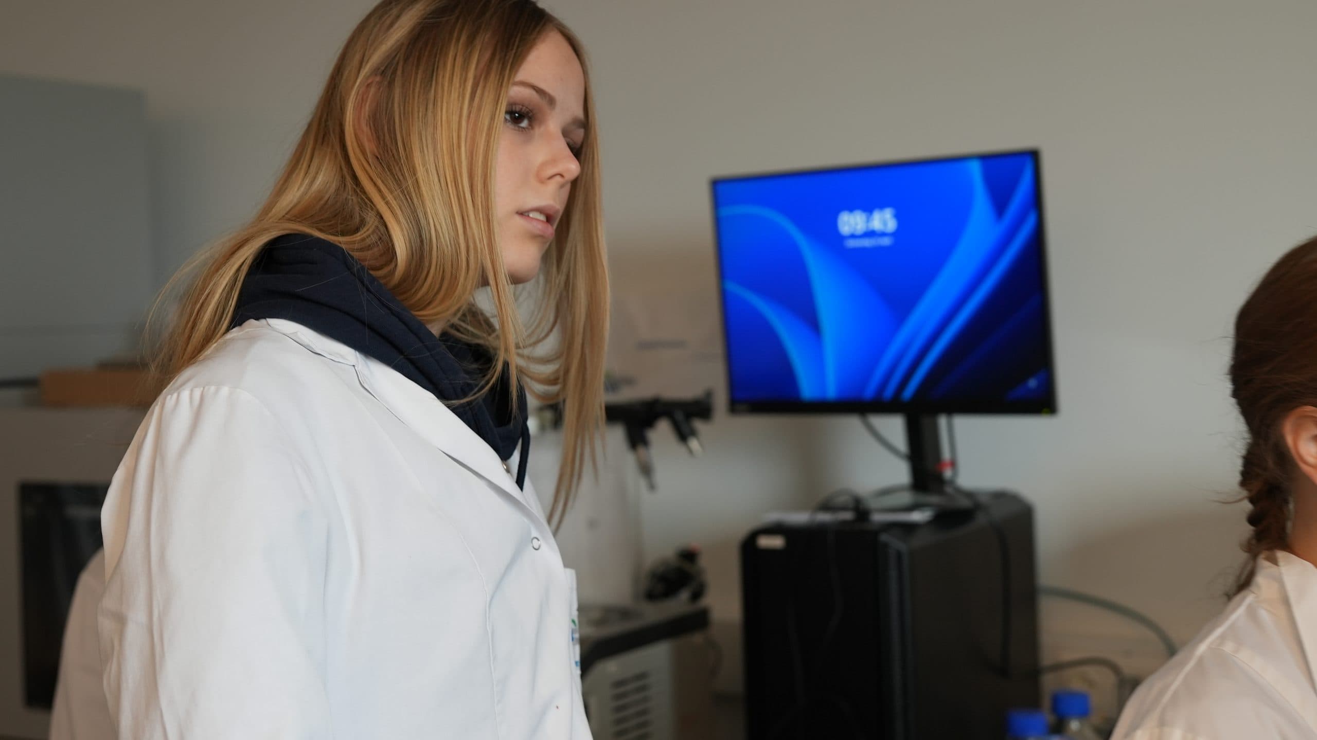 Eine junge Frau in einem weißen Laborkittel steht in einem Labor. Im Hintergrund befinden sich wissenschaftliche Geräte und ein Computermonitor, der die Uhrzeit 09:45 anzeigt. Eine weitere Person ist teilweise rechts zu sehen.