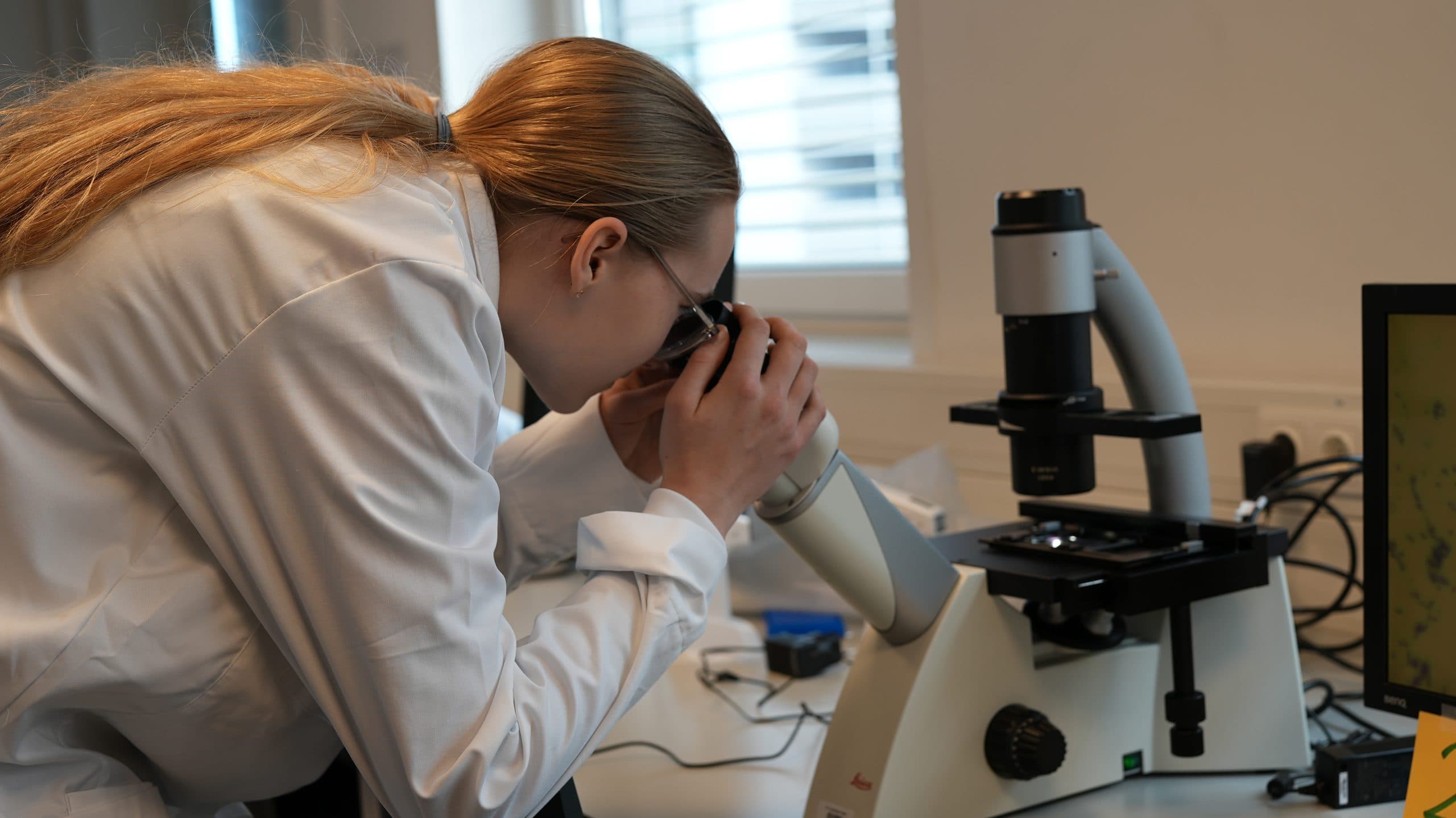 Eine Person in einem weißen Laborkittel mit zurückgebundenen langen Haaren blickt in einer Laborumgebung in ein Mikroskop, während auf dem Schreibtisch daneben ein Computermonitor und wissenschaftliche Geräte zu sehen sind.