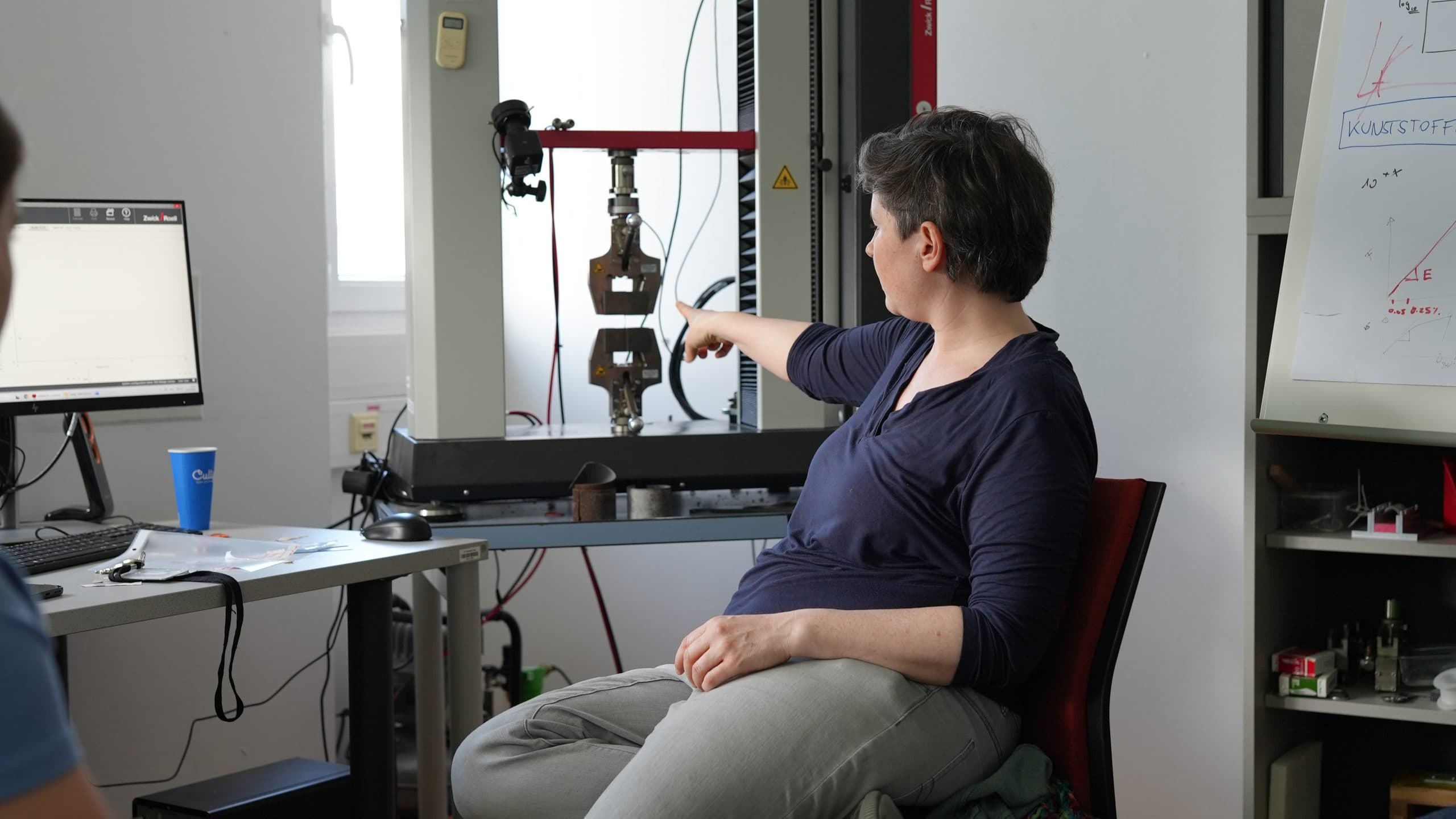 Eine in einem Büro sitzende Person zeigt auf Geräte an einer Testmaschine, während in der Nähe Computerbildschirme und Tabellen zu sehen sind. Die Umgebung erinnert an ein wissenschaftliches oder technisches Labor.