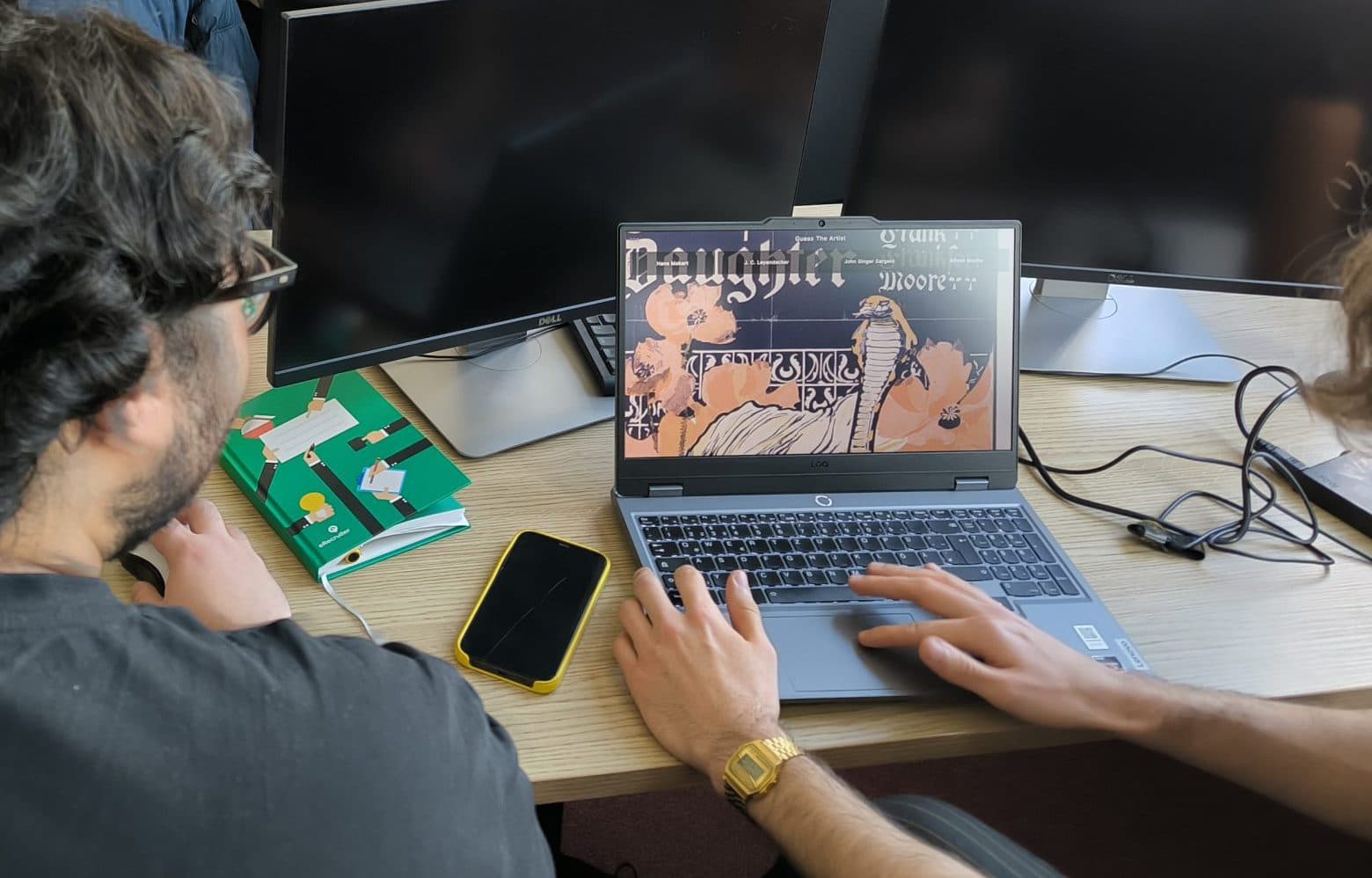 Eine Gruppe von Personen sitzt an Tischen mit mehreren Computermonitoren und Laptops und arbeitet in einer Art Klassenzimmer zusammen. Auf einigen Bildschirmen wird Code angezeigt, während auf einem ein Hintergrundbild mit Anime-Themen zu sehen ist.