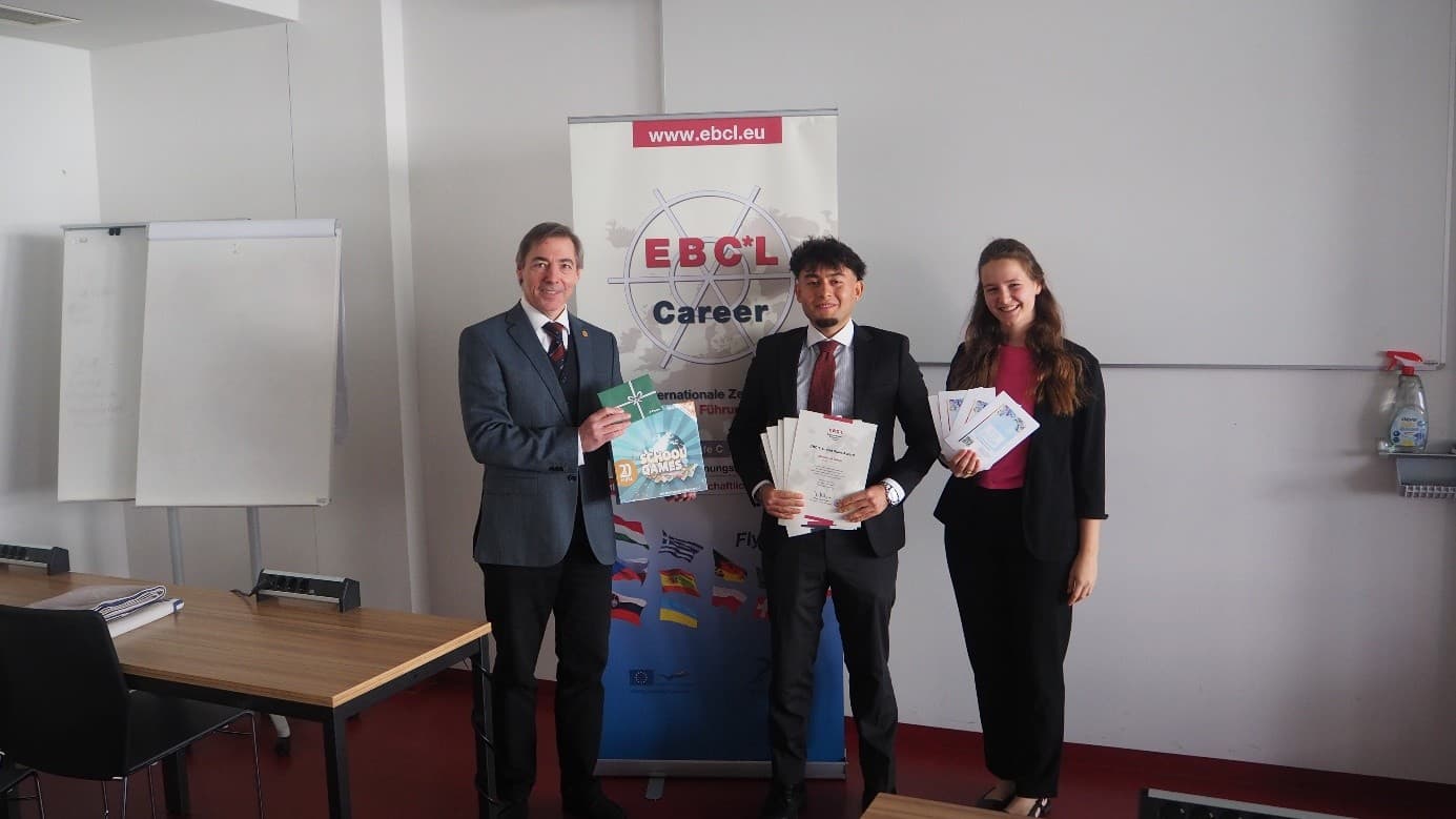 Drei Personen in Geschäftskleidung stehen in einem Raum, lächeln und halten Zertifikate vor einem EBCL-Karrierebanner. Im Raum sind zwei Tische, ein Whiteboard und Regale mit Reinigungsmitteln zu sehen.