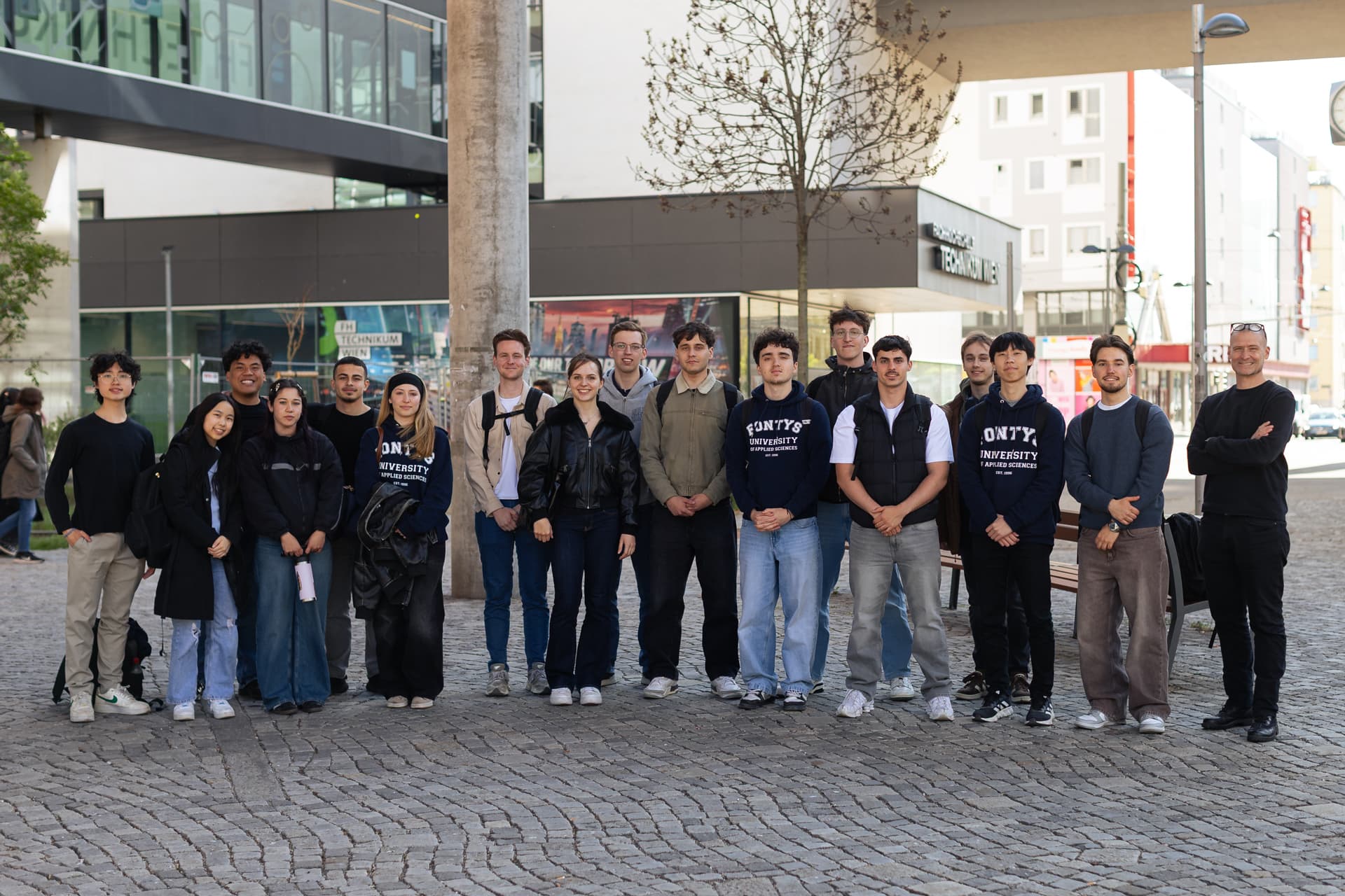 Eine Gruppe von 17 jungen Erwachsenen und ein älterer Erwachsener stehen gemeinsam im Freien auf einem Kopfsteinpflaster in einer Stadt und posieren für ein Gruppenfoto. Einige tragen dunkle Sweatshirts der ONYX University; im Hintergrund sind Gebäude zu sehen.