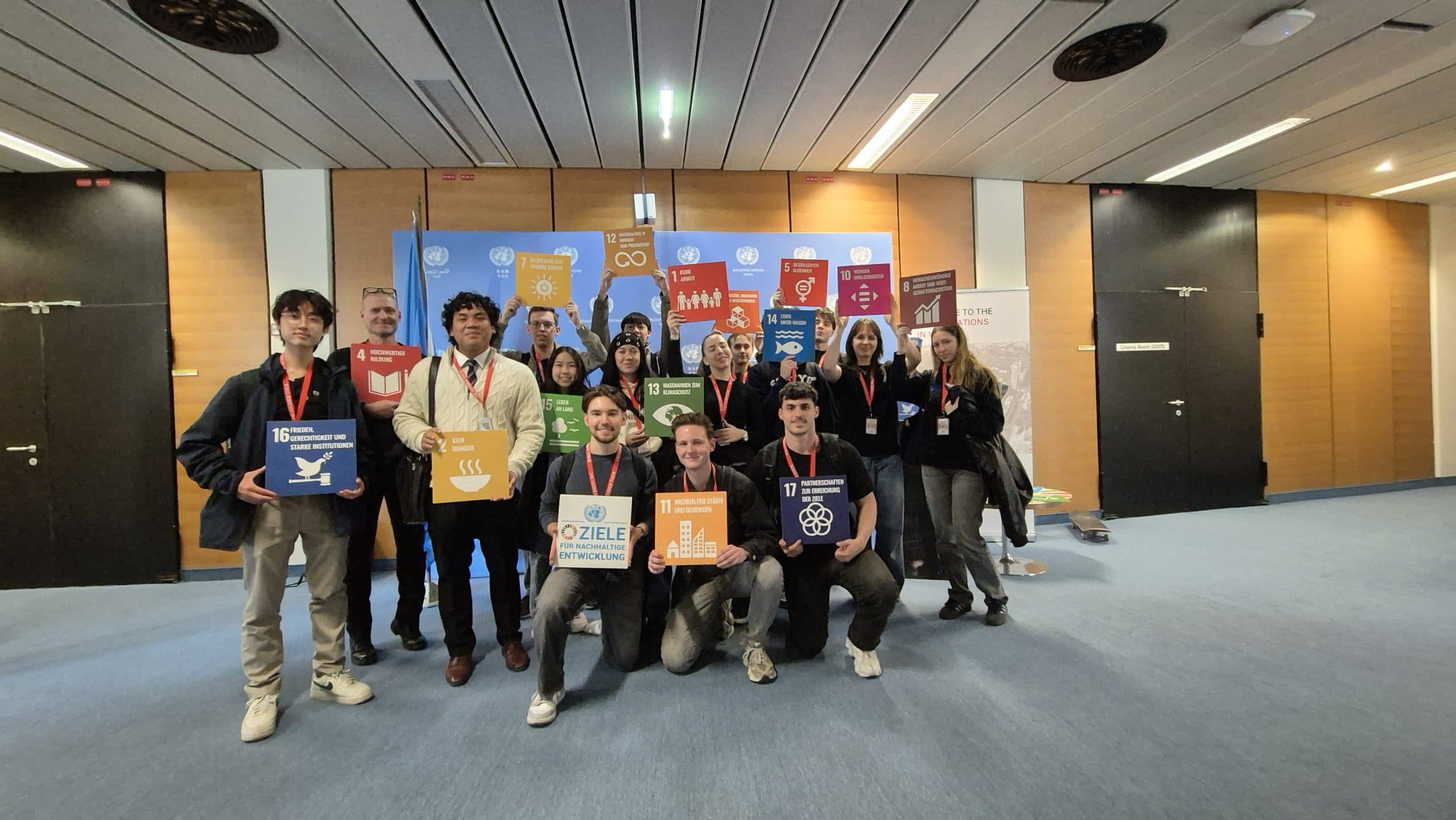 Eine Gruppe von Menschen steht drinnen und hält bunte Schilder, die verschiedene Ziele für nachhaltige Entwicklung der Vereinten Nationen repräsentieren, und lächelt in die Kamera vor einem Display mit entsprechenden Symbolen und Logos.