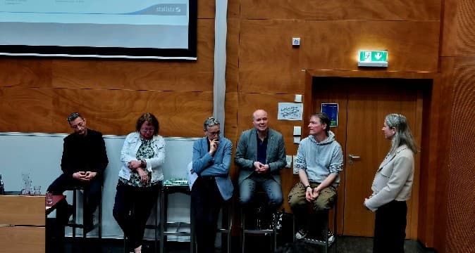 Sechs Personen sind im vorderen Teil eines Hörsaals versammelt, fünf davon sitzen auf Hockern, eine steht. Sie scheinen in eine Diskussion vertieft zu sein. Hinter ihnen befinden sich eine holzgetäfelte Wand und eine Projektionsfläche.