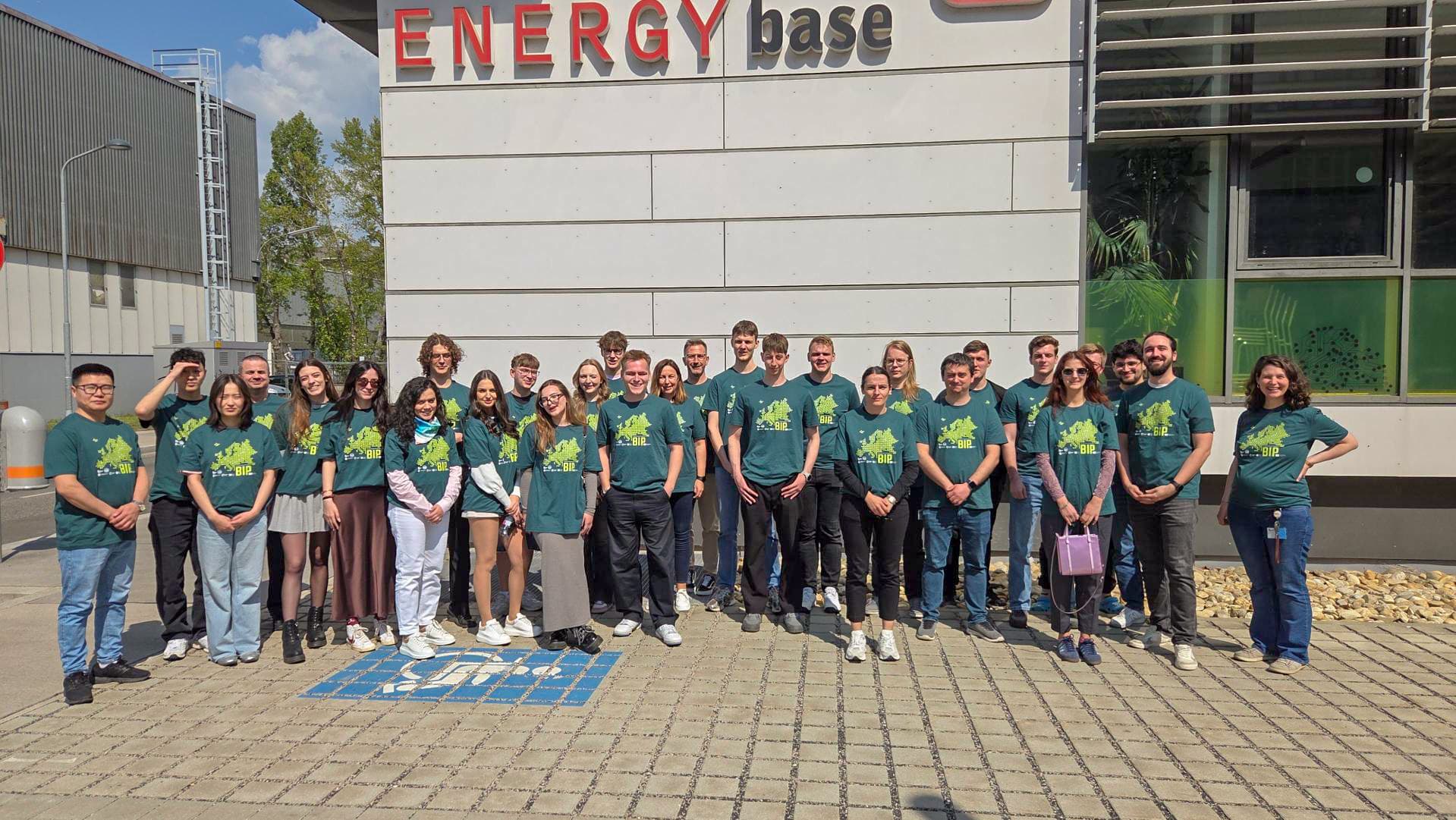 Eine Gruppe von etwa 30 Personen in passenden grünen T-Shirts posiert an einem sonnigen Tag vor einem Gebäude mit dem Schild ENERGY base. Sie stehen auf einer gepflasterten Fläche und lächeln in die Kamera.