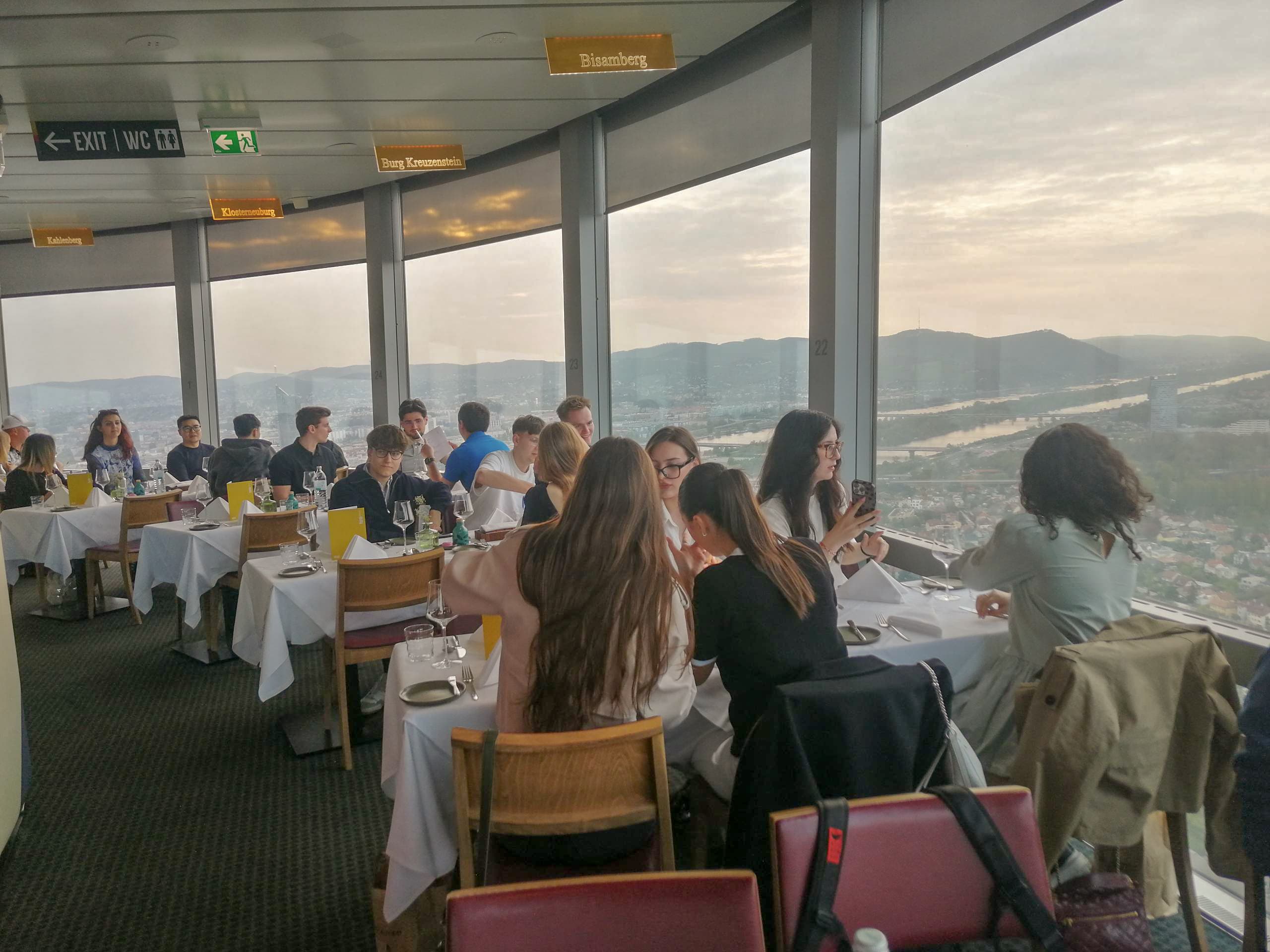 Die Menschen sitzen an Tischen in einem Restaurant mit großen Fenstern, die einen Panoramablick auf einen Fluss, die Stadt und die fernen Hügel bei Sonnenuntergang bieten. Die Atmosphäre ist gesellig und entspannt, das natürliche Licht erfüllt den Raum.