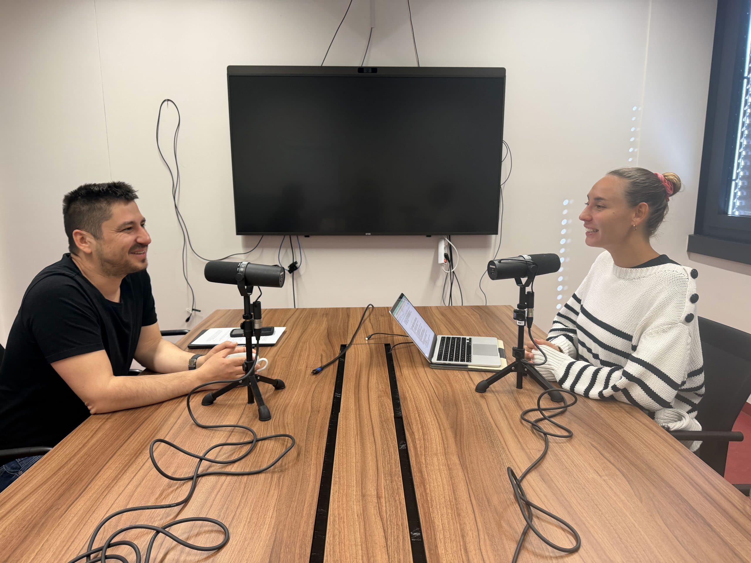 Zwei Personen sitzen sich an einem Tisch mit Mikrofonen, einem Laptop und Notizen gegenüber und nehmen einen Podcast oder ein Interview in einem modernen Raum mit einem großen Fernsehbildschirm an der Wand hinter ihnen auf.