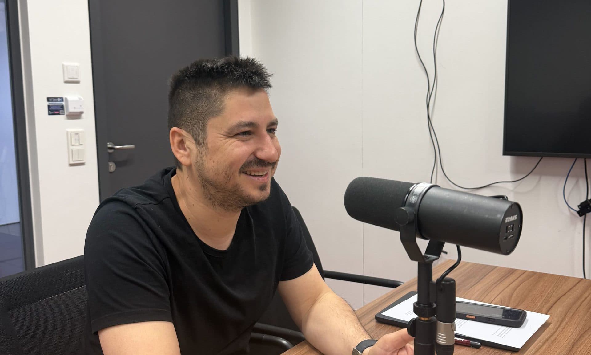 Ein Mann mit kurzen dunklen Haaren und einem Bart, der ein schwarzes T-Shirt trägt, sitzt an einem Holztisch und spricht in einem modernen Büro in ein Mikrofon. Er lächelt, und auf dem Tisch neben ihm liegen ein Notizblock und ein Telefon.
