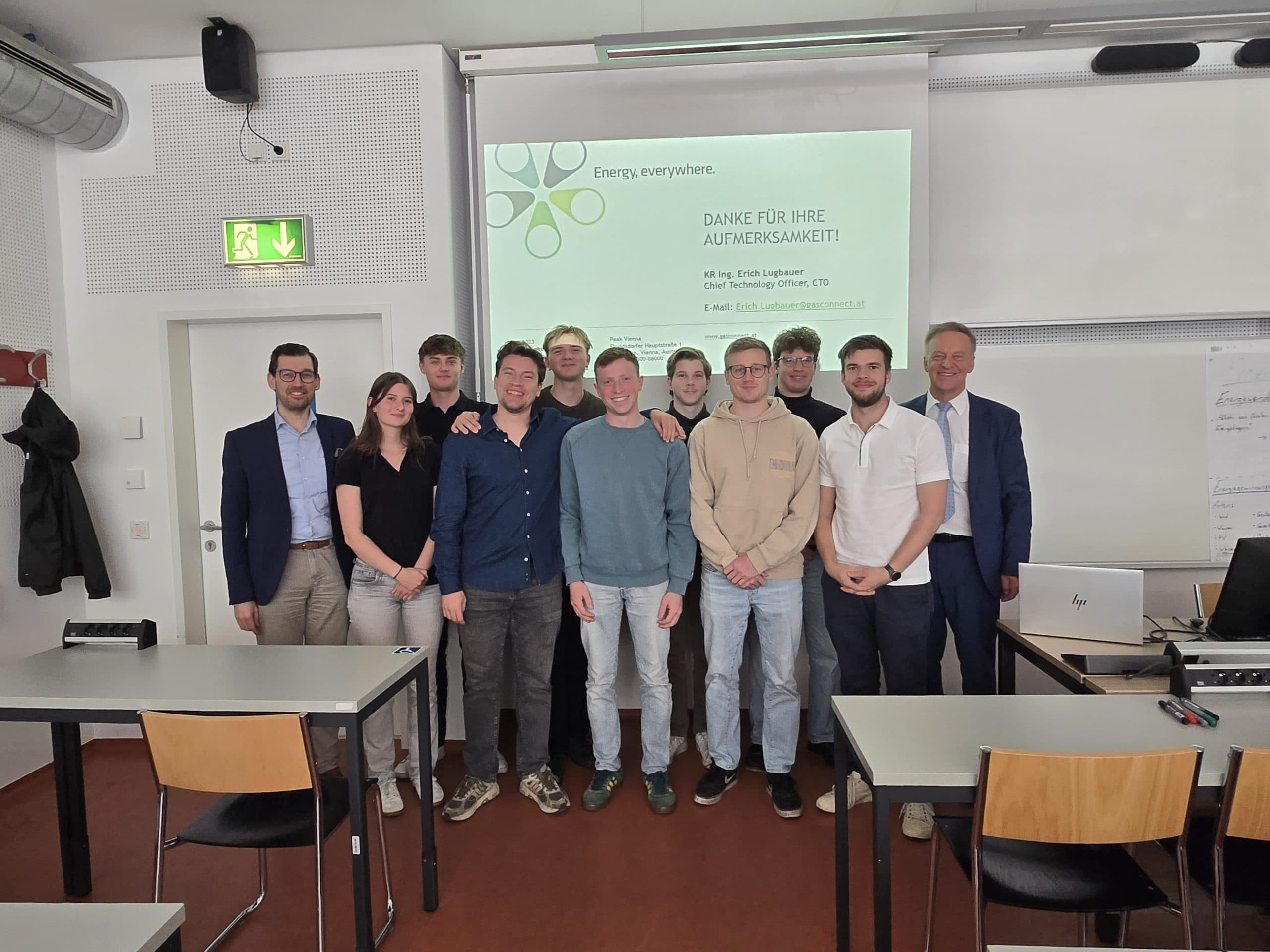 Eine Gruppe von zehn Personen steht und lächelt für ein Foto in einem Klassenzimmer. Hinter ihnen ist eine projizierte Präsentationsfolie mit dem Titel "Energie, überall" zu sehen, und im Vordergrund stehen Tische mit Stühlen.