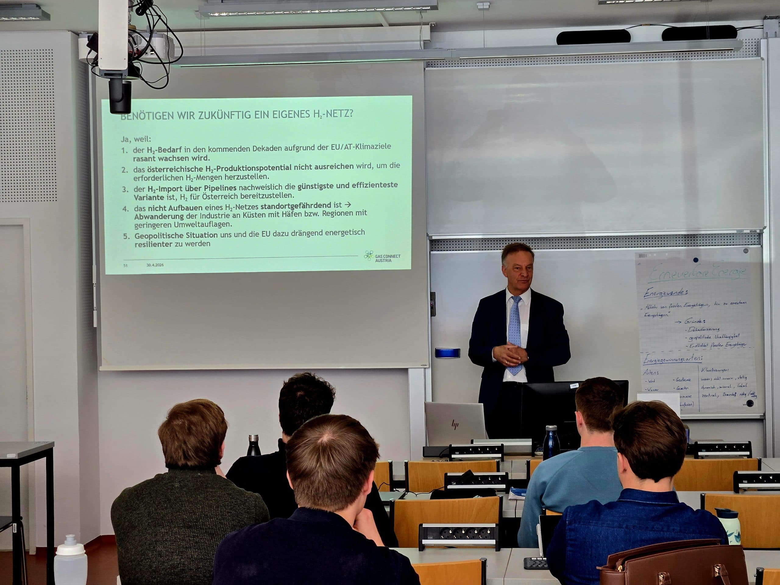Ein Mann im Anzug hält vor einer Gruppe von Schülern eine Diashow. Auf dem Bildschirm wird ein deutscher Text angezeigt, und auf einem Flipchart neben ihm werden Notizen gemacht. Mehrere Schüler sitzen mit dem Gesicht zum Vortragenden und machen sich Notizen.