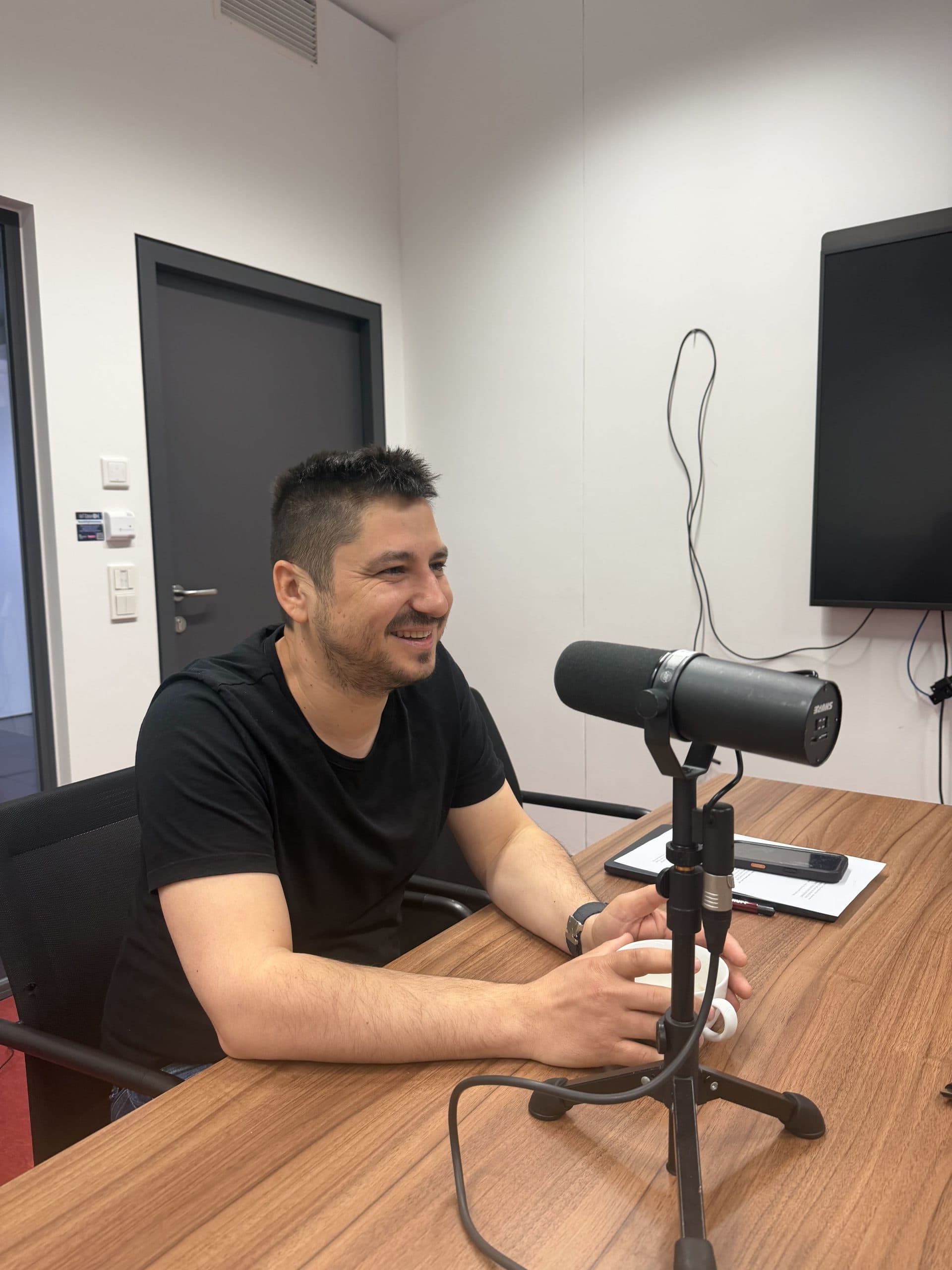 Ein Mann mit kurzen dunklen Haaren und einem Bart, der ein schwarzes T-Shirt trägt, sitzt an einem Holztisch mit einem Mikrofon, einem Notebook und einem Smartphone und lächelt während einer Podcast-Aufnahme in einem modernen Büro.