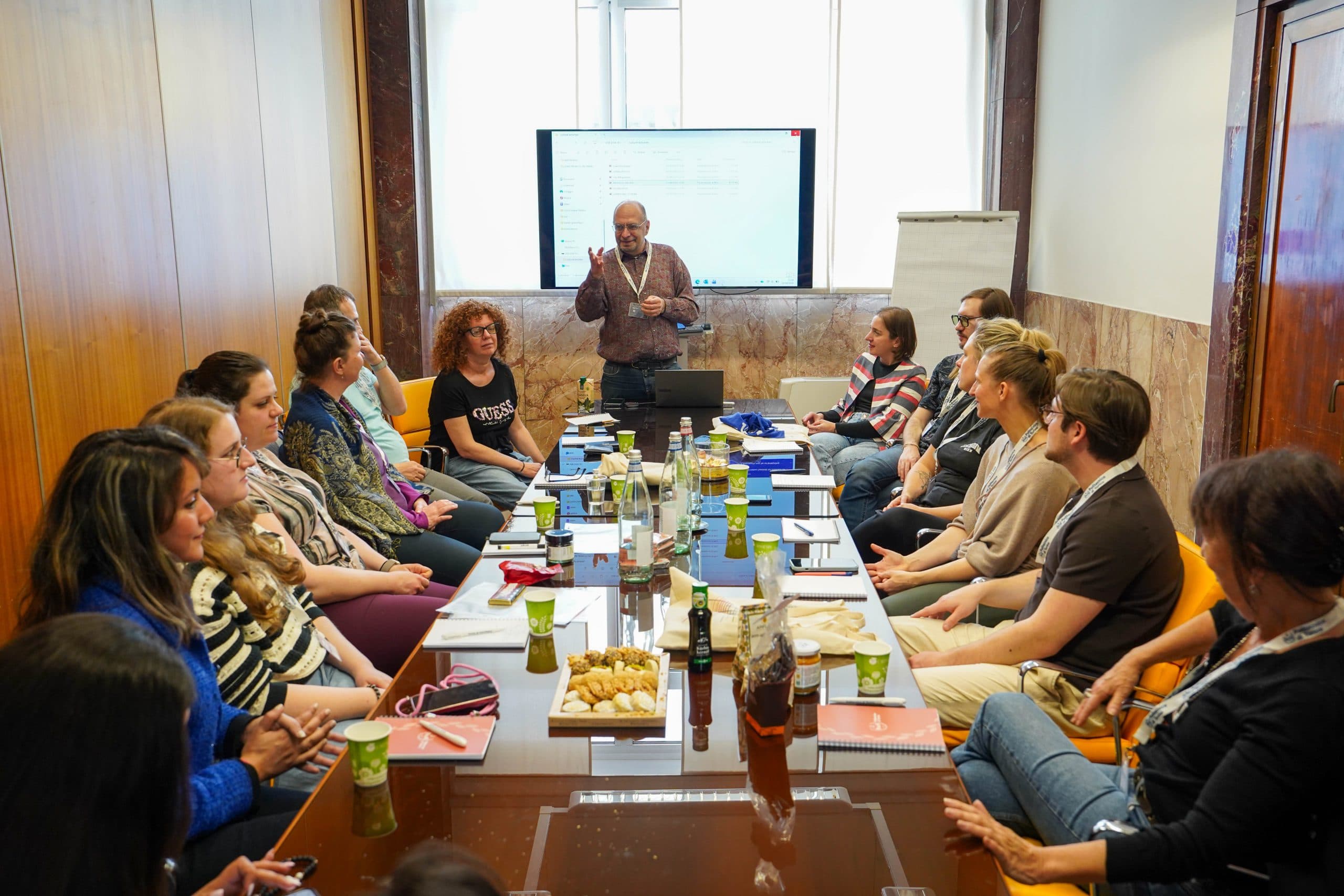 Eine Gruppe von Personen sitzt um einen Konferenztisch mit Getränken und Snacks und hört einem Mann zu, der am vorderen Ende des Raumes neben einem großen Bildschirm und einem Flipchart steht und spricht.