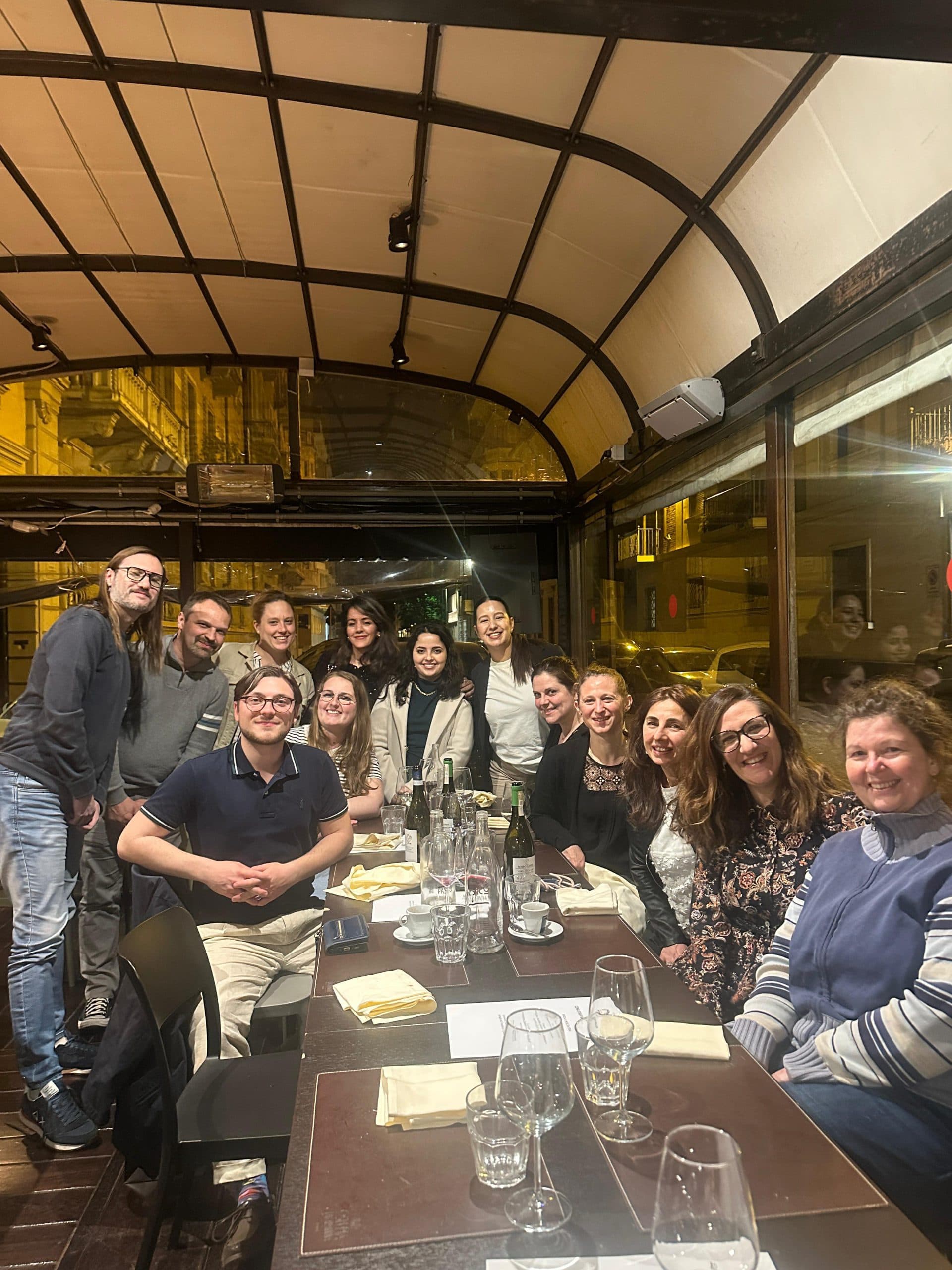Eine Gruppe von dreizehn Personen, die lächelnd um einen langen Tisch in einem Restaurant mit leeren Tellern, Gläsern und Weinflaschen auf dem Tisch posieren, in einem warm beleuchteten Raum mit großen Fenstern.