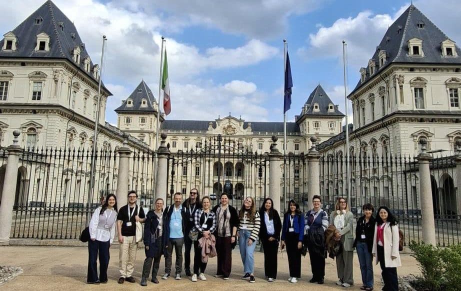 Eine Gruppe von Menschen steht vor einem großen historischen Gebäude mit kunstvoller Architektur, eisernen Toren und beflaggten Masten unter einem teilweise bewölkten Himmel. Die Gruppe scheint für ein Gruppenfoto zu posieren.