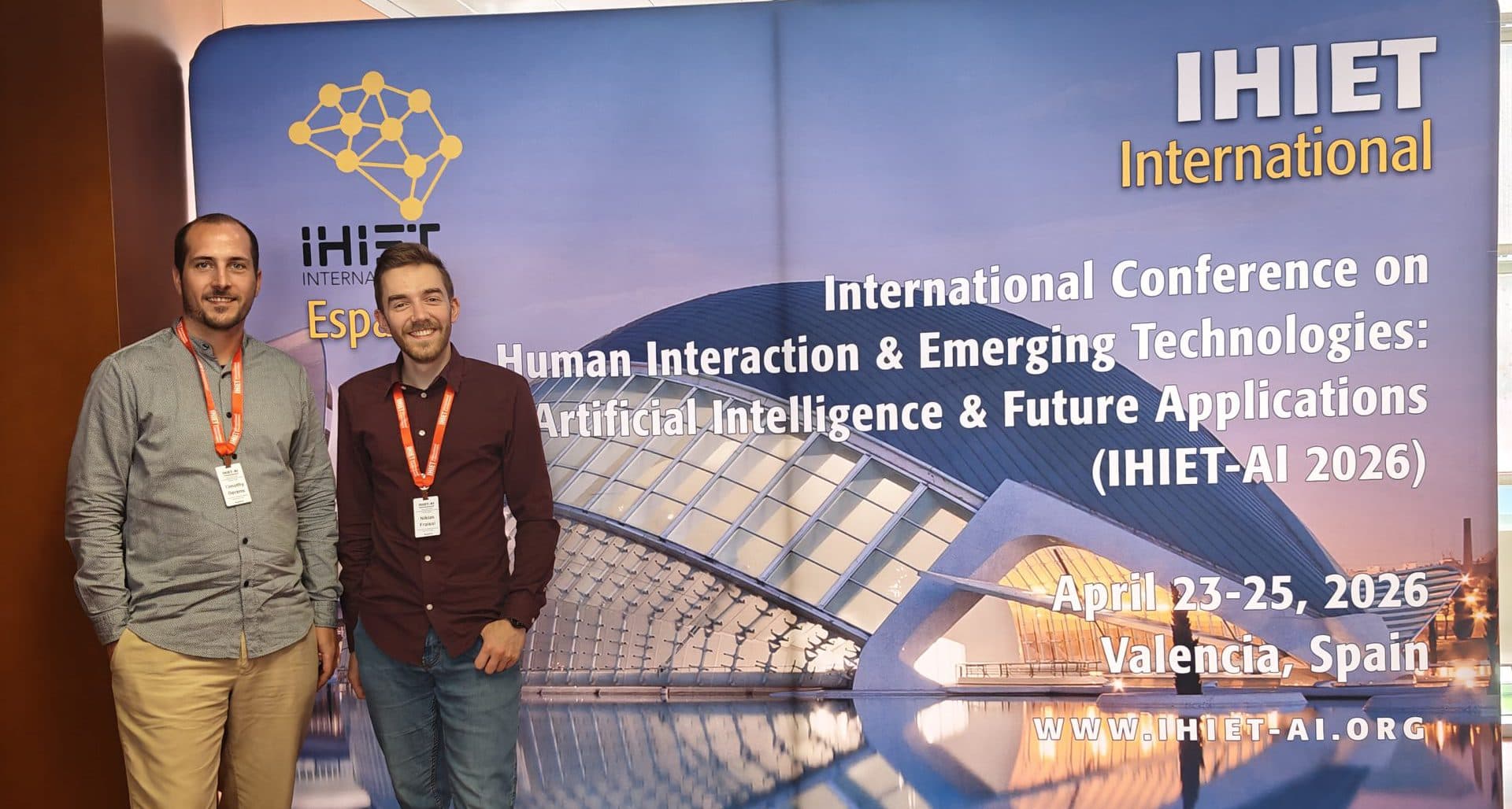 Zwei Männer stehen lächelnd vor einem Konferenzbanner für die International Conference on Human Interaction & Emerging Technologies (IHIET-AI 2026) in Valencia, Spanien. Auf dem Banner sind Veranstaltungsdetails und ein modernes Gebäude abgebildet.