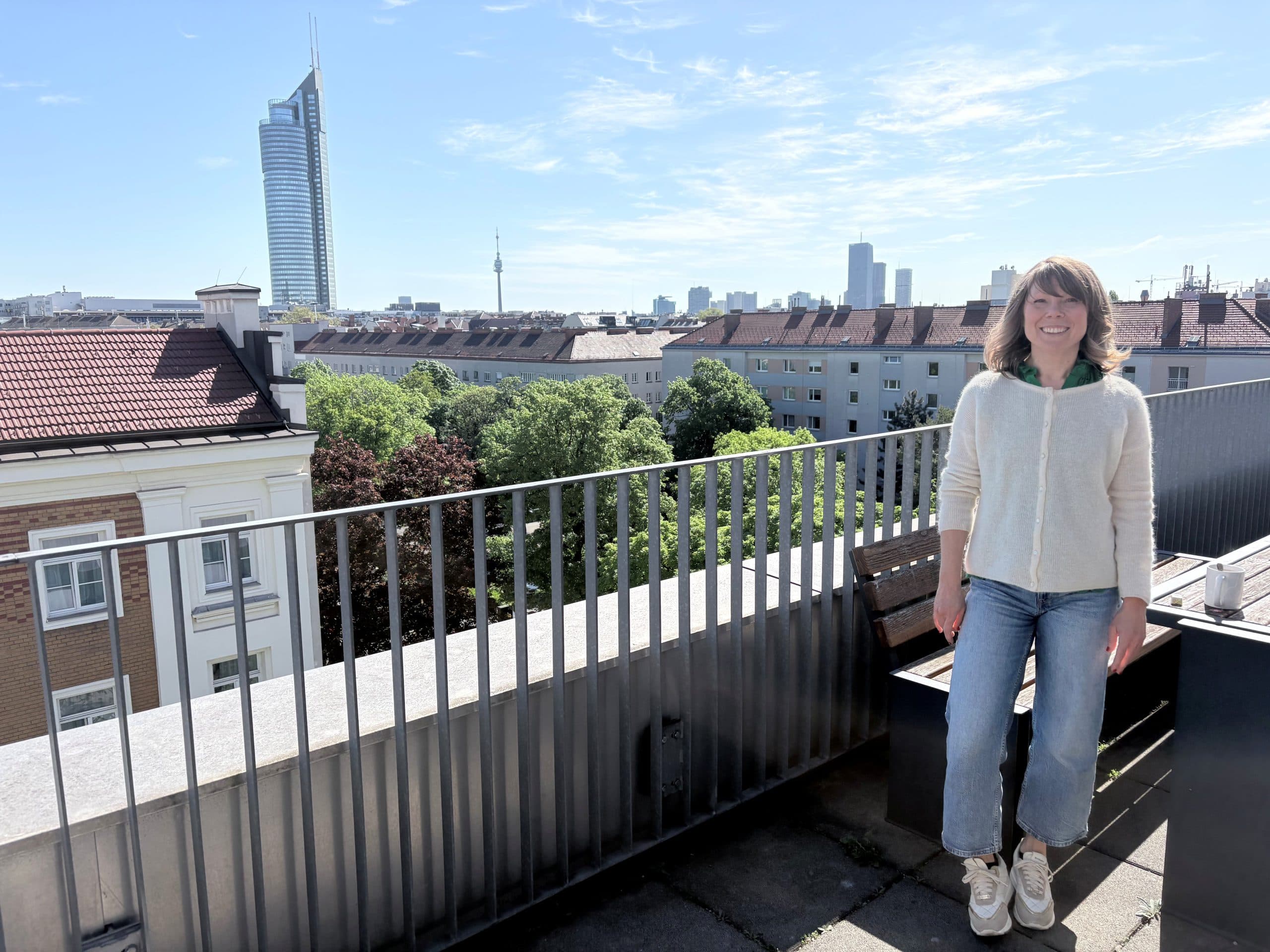 Eine Frau mit schulterlangem Haar, die einen hellen Pullover und Jeans trägt, sitzt an einem sonnigen Tag auf einer Bank auf einer Dachterrasse mit der Skyline der Stadt, Bäumen und hohen modernen Gebäuden im Hintergrund.