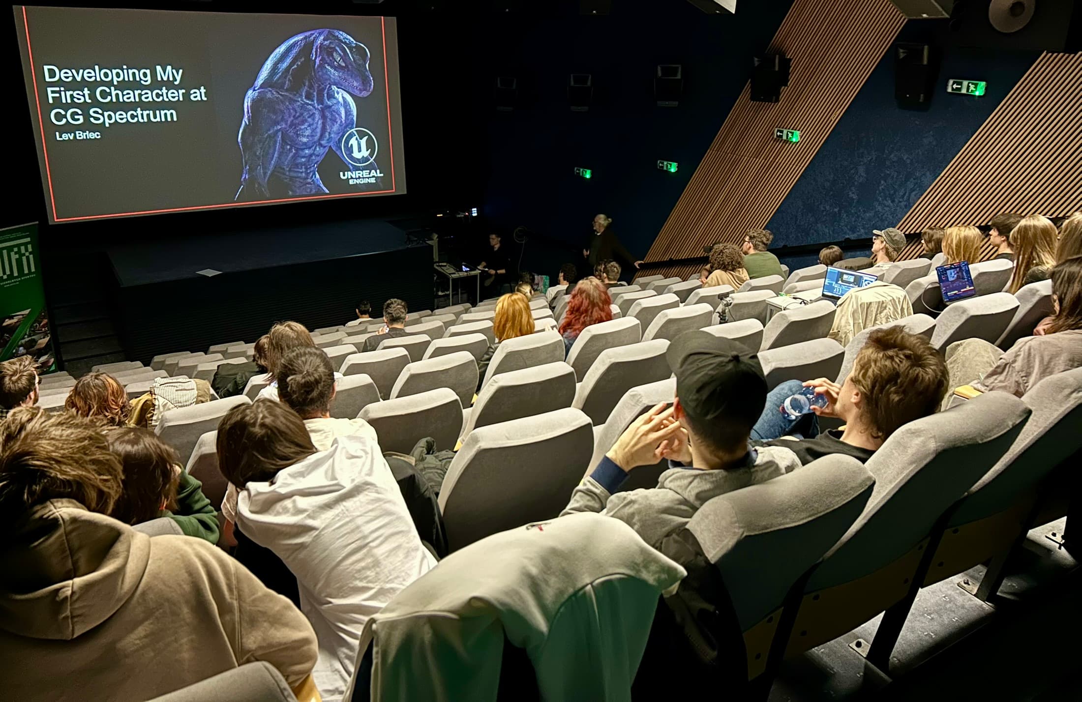 Ein Publikum in einem Vortragssaal sieht sich eine Präsentation mit dem Titel 'Developing My First Character at CG Spectrum' an, bei der ein Dinosaurierbild und ein Unreal-Engine-Logo auf einer großen Leinwand im Vordergrund zu sehen sind.