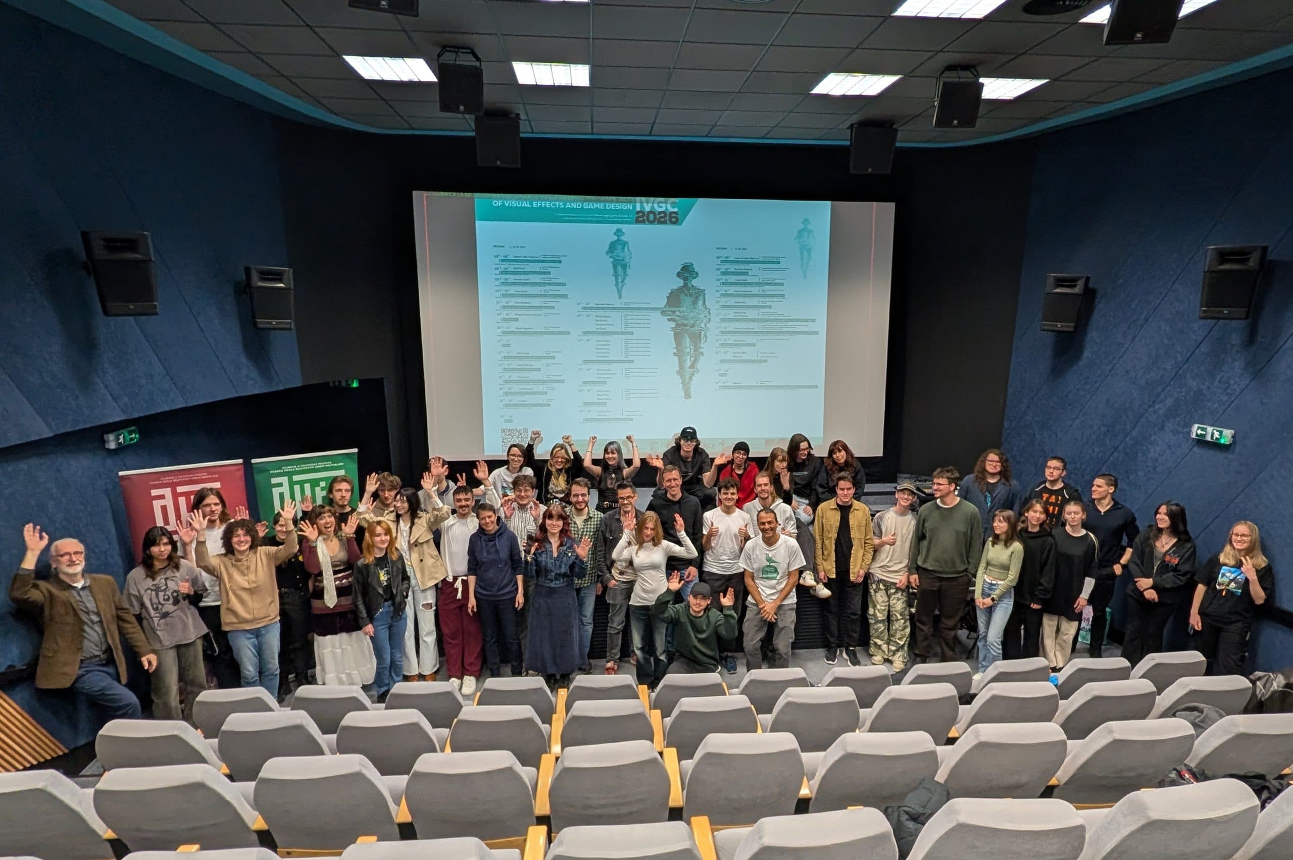 Eine Gruppe von Menschen steht am Eingang eines Kinos, lächelt und winkt in die Kamera. Hinter ihnen befindet sich ein großer Bildschirm, auf dem ein Programmablaufplan angezeigt wird. Die Sitze im Vordergrund sind größtenteils leer.