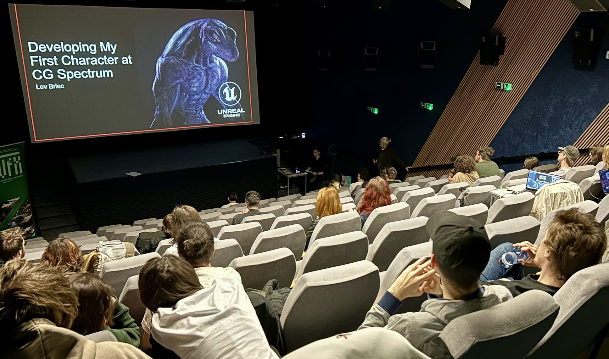 Das Publikum sitzt in einem Theater und sieht sich eine Präsentation mit dem Titel Developing My First Character at CG Spectrum an, bei der auf einer großen Leinwand im vorderen Teil des Raums ein Dinosaurier abgebildet ist.