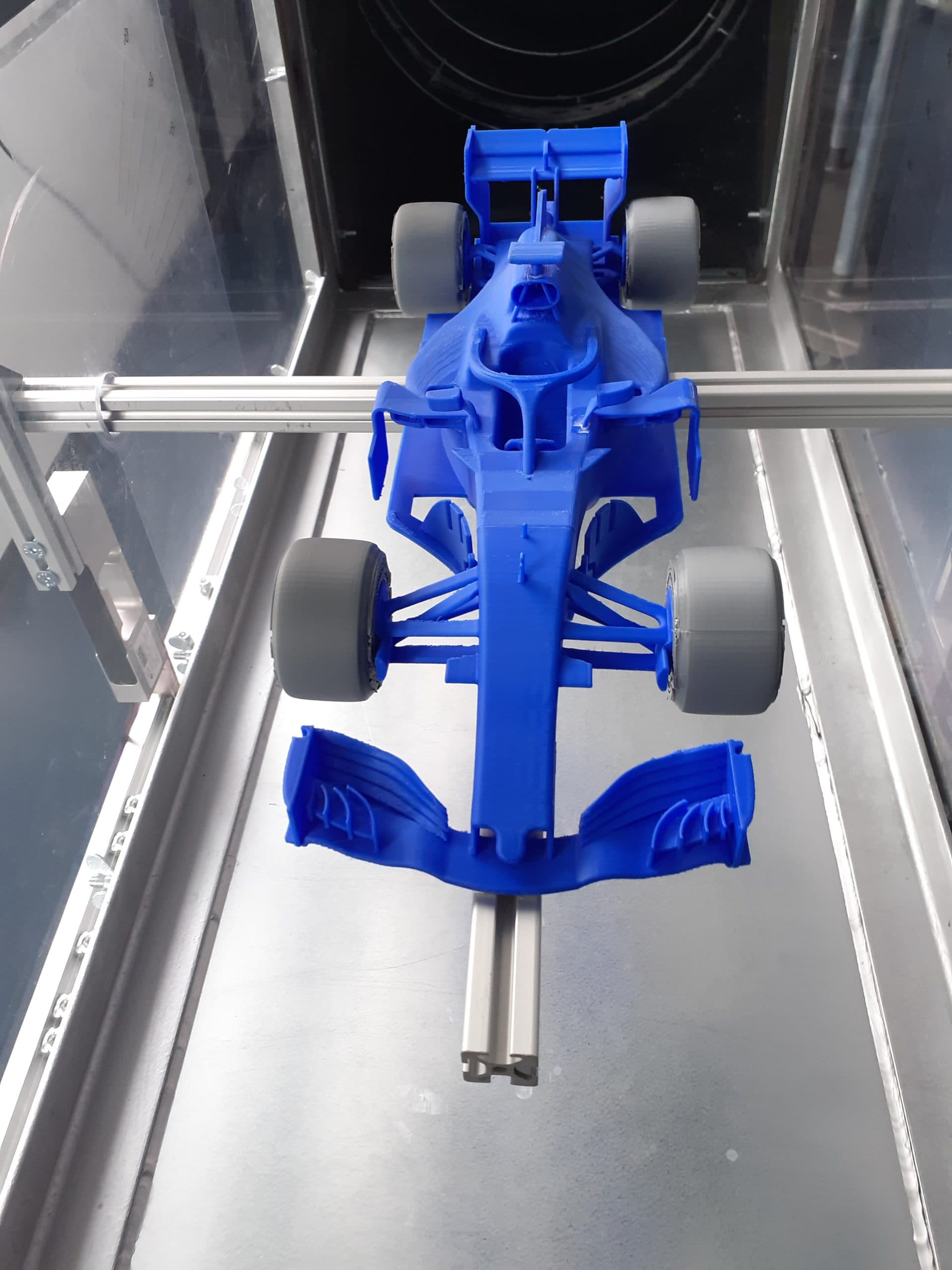 Ein blaues 3D-gedrucktes Modell eines Formel-1-Rennwagens wird in einem 3D-Drucker ausgestellt, von vorne betrachtet mit sichtbaren Rädern, Spoiler und aerodynamischen Details.