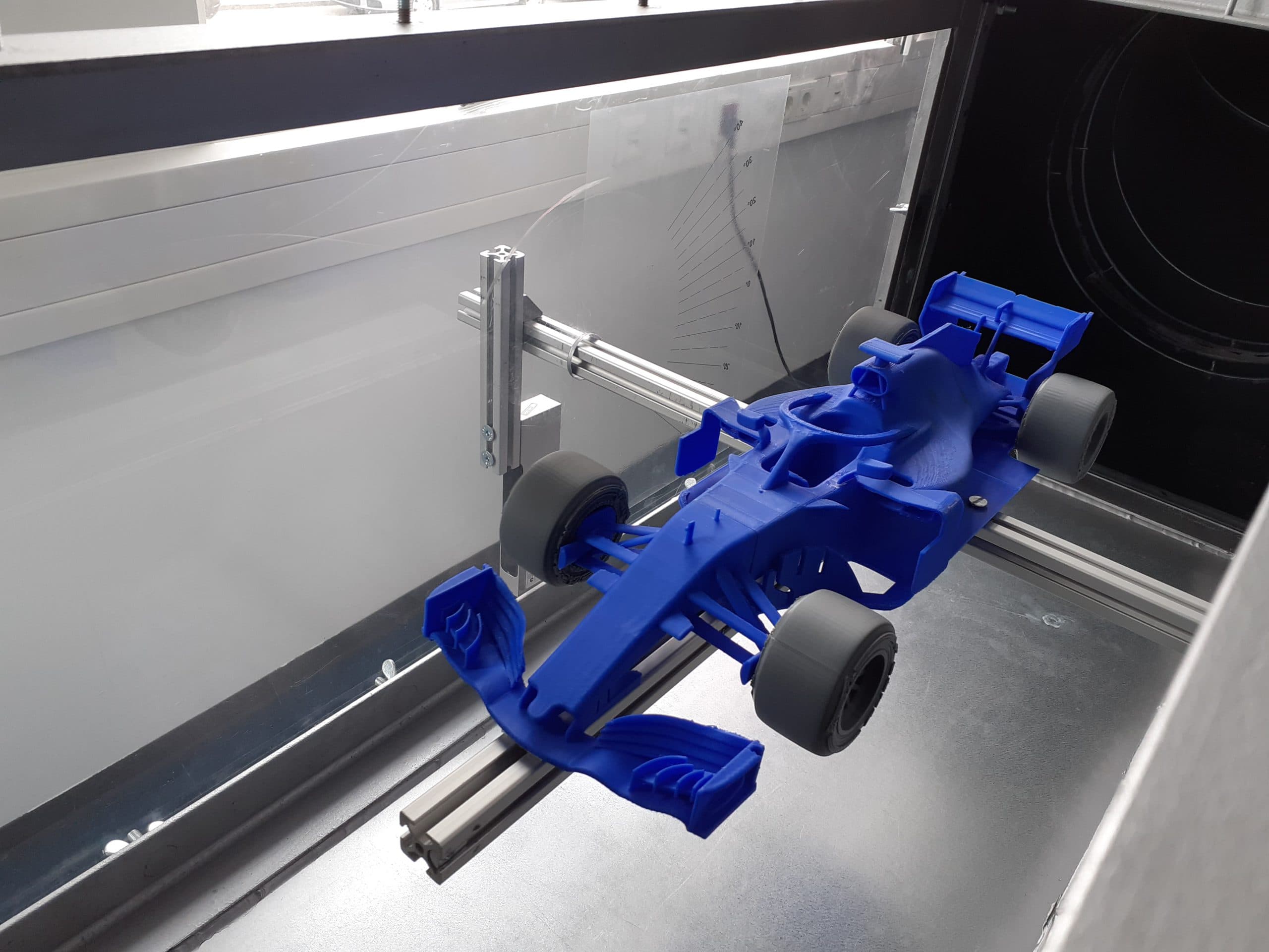 Ein blaues Modell eines Formel-1-Autos ist auf einer Metallschiene in einem Windkanal montiert, wahrscheinlich für aerodynamische Tests. Im Hintergrund sind Messmarkierungen und Steckdosen zu sehen.