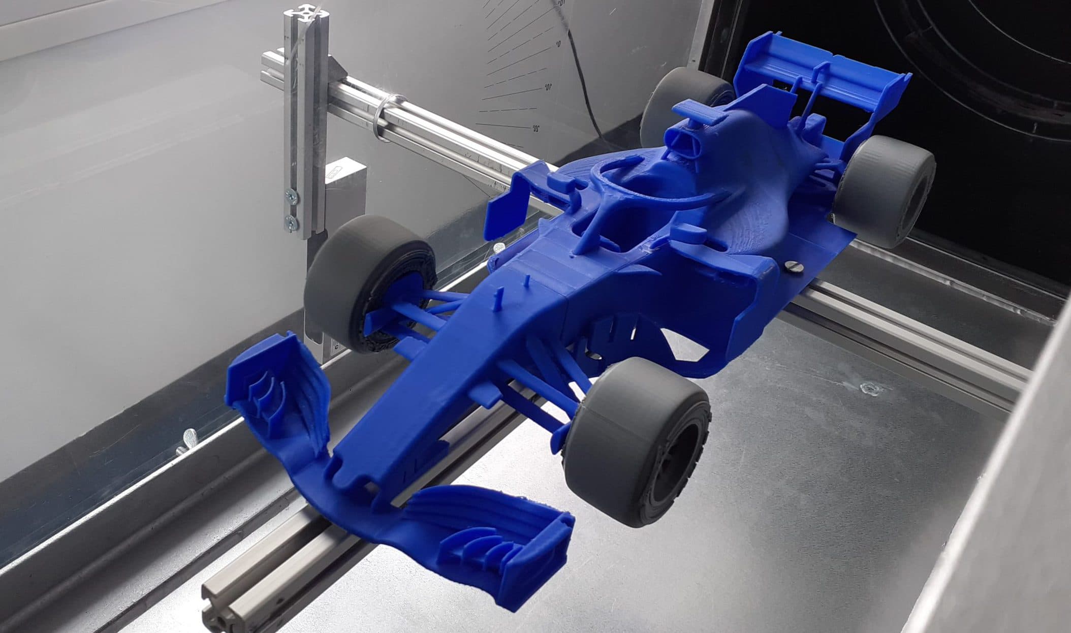 Ein blaues Modell eines Formel-1-Rennwagens ist an einer Metallstange in einem Windkanal befestigt, der wahrscheinlich für aerodynamische Tests verwendet wird. Im Hintergrund sind ein Fenster und Messmarkierungen zu sehen.