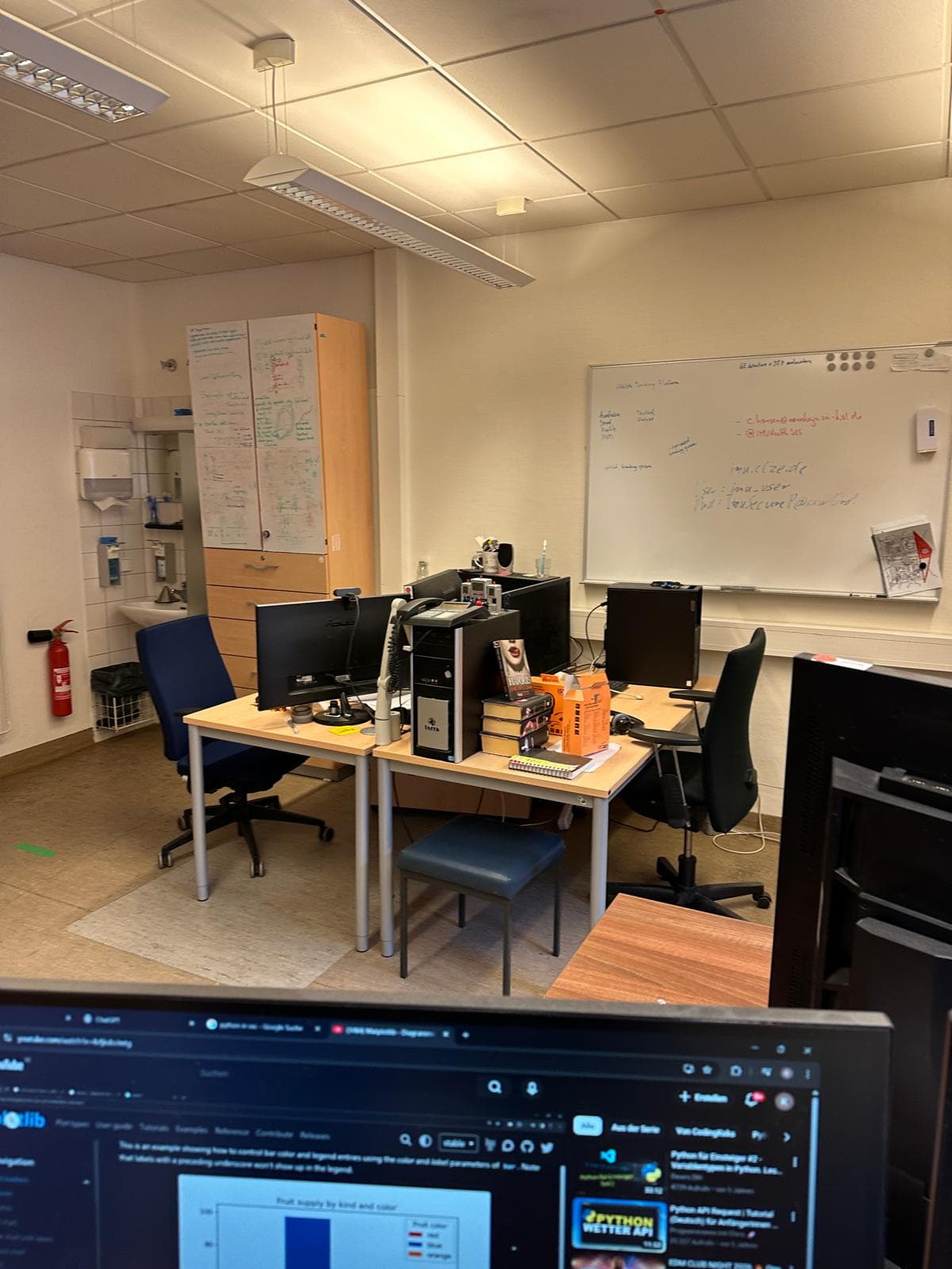 Ein kleines Büro mit Schreibtischen, Computern, Stühlen und Monitoren. An den Wänden sind Whiteboards mit Notizen und Diagrammen angebracht, und auf einem Schreibtisch stehen Snacks und Getränke. Ein Feuerlöscher steht in der Ecke neben einem Schrank.
