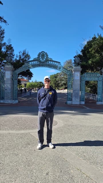Eine Person mit Sonnenbrille, weißer Mütze und blauer Jacke steht an einem sonnigen Tag lächelnd vor dem dekorativen Sather Gate der UC Berkeley. Im Hintergrund sind Bäume und ein paar Menschen zu sehen.