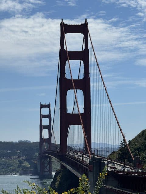 Die Golden Gate Bridge in San Francisco an einem sonnigen Tag, mit blauem Himmel und vereinzelten Wolken darüber und Autos, die die Brücke über das Wasser überqueren. Im Hintergrund sind grüne Hügel und das Stadtbild zu sehen.