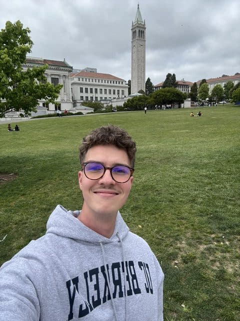 Eine Person, die eine Brille und einen grauen UC BERKELEY-Kapuzenpulli trägt, lächelt, während sie ein Selfie auf einer großen Wiese mit dem UC Berkeley-Campus und dem Campanile-Turm im Hintergrund unter einem bewölkten Himmel macht.