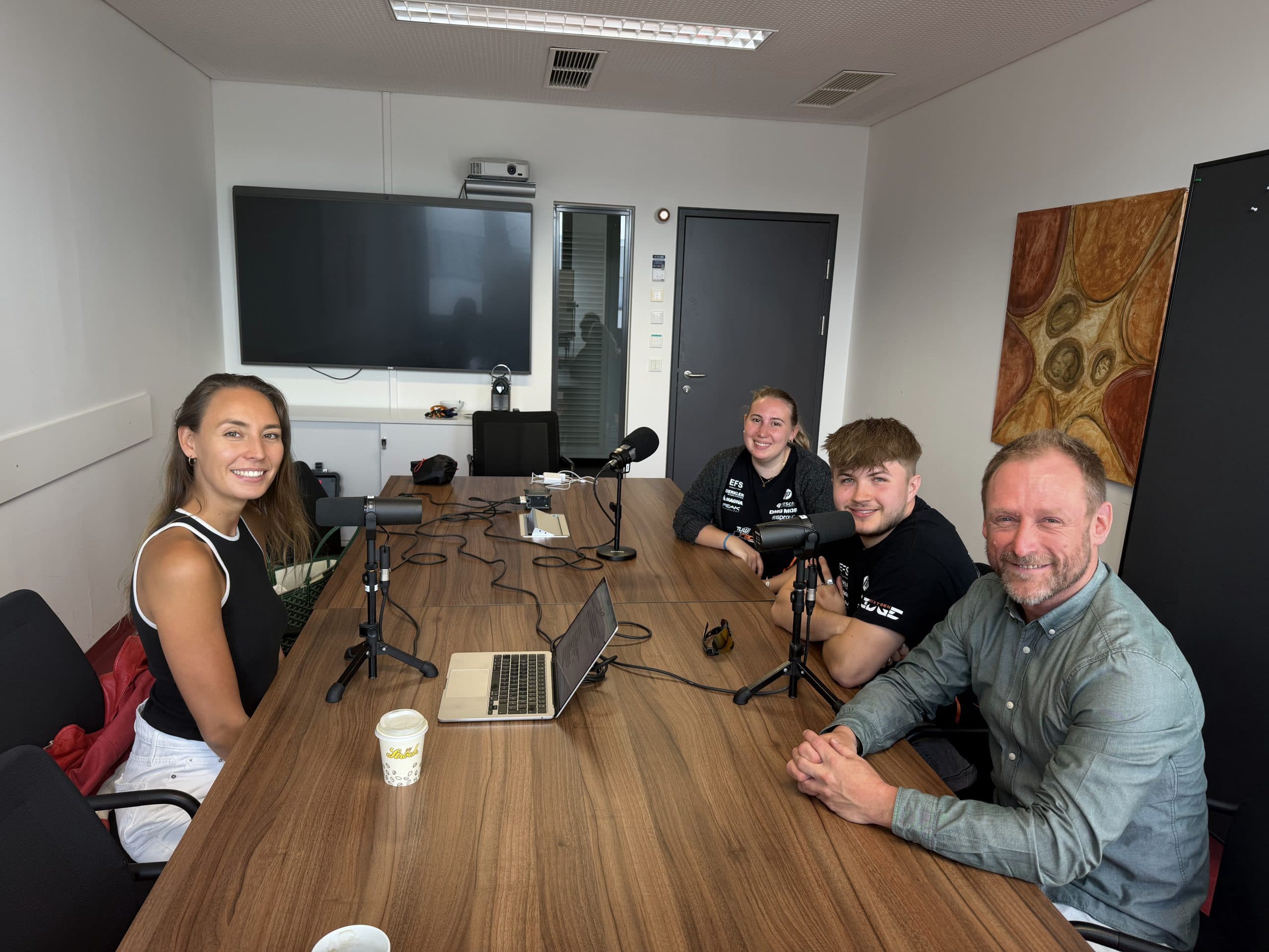 Vier Personen sitzen mit Mikrofonen, Laptops und Kaffeetassen um einen Tisch herum und scheinen einen Podcast oder ein Interview in einem modernen Besprechungsraum mit einem großen Fernseher und Wandkunst im Hintergrund aufzunehmen.